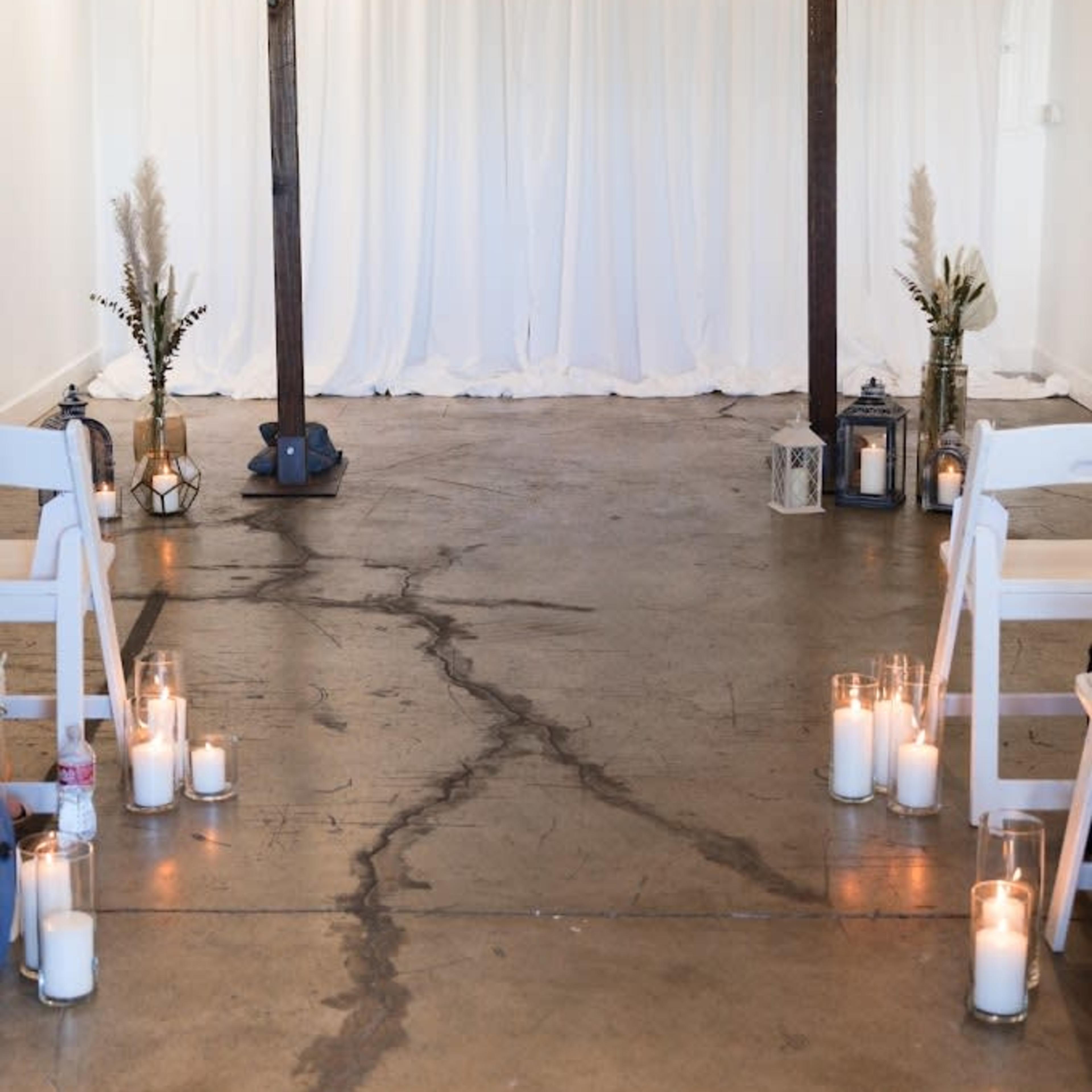 A minimalist wedding altar with a wooden arch, surrounded by white chairs and lit candles on a concrete floor beneath a white backdrop.