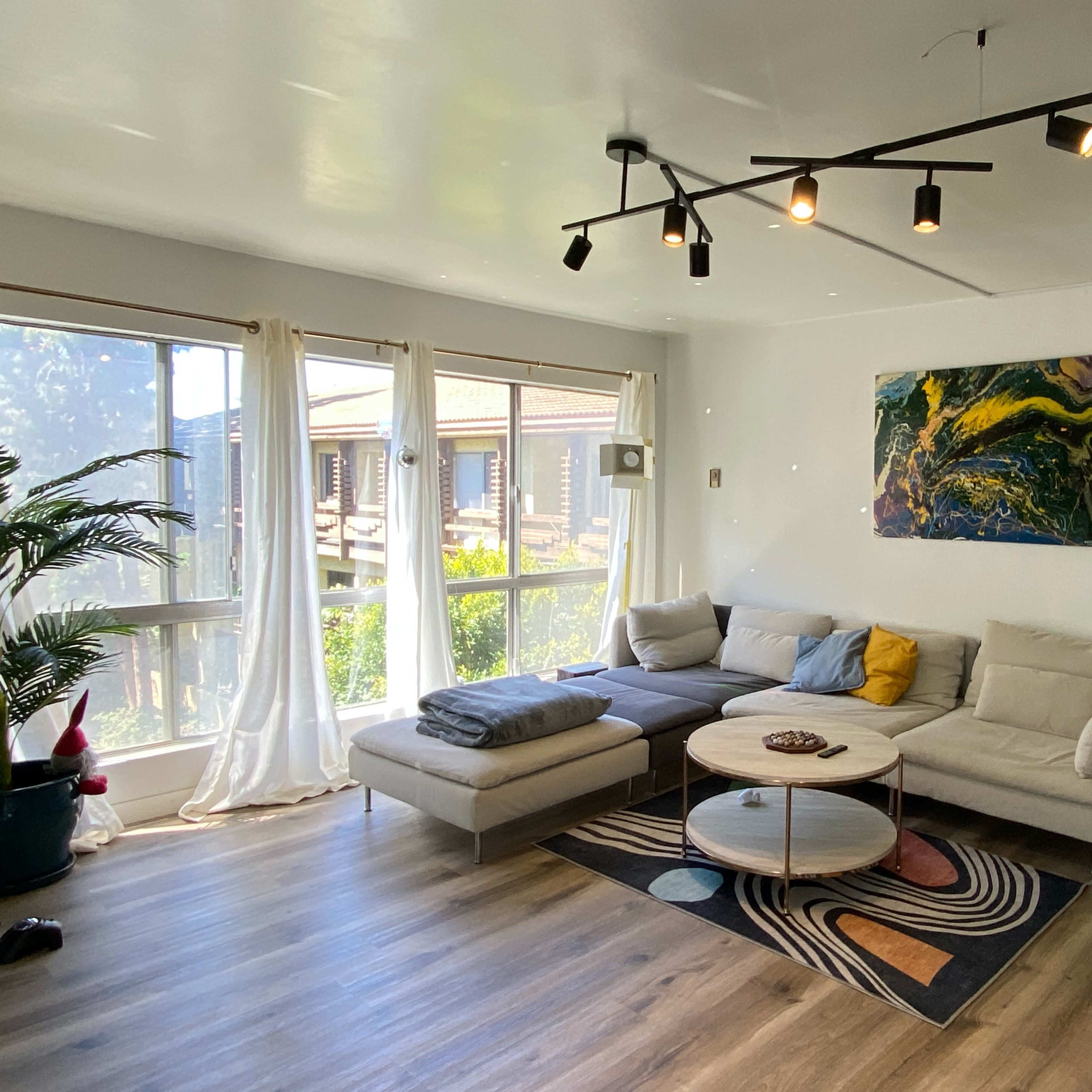 The living room features a light-colored sectional sofa, a round coffee table, and large windows that let in natural light, with plants and decorative artwork enhancing the space.