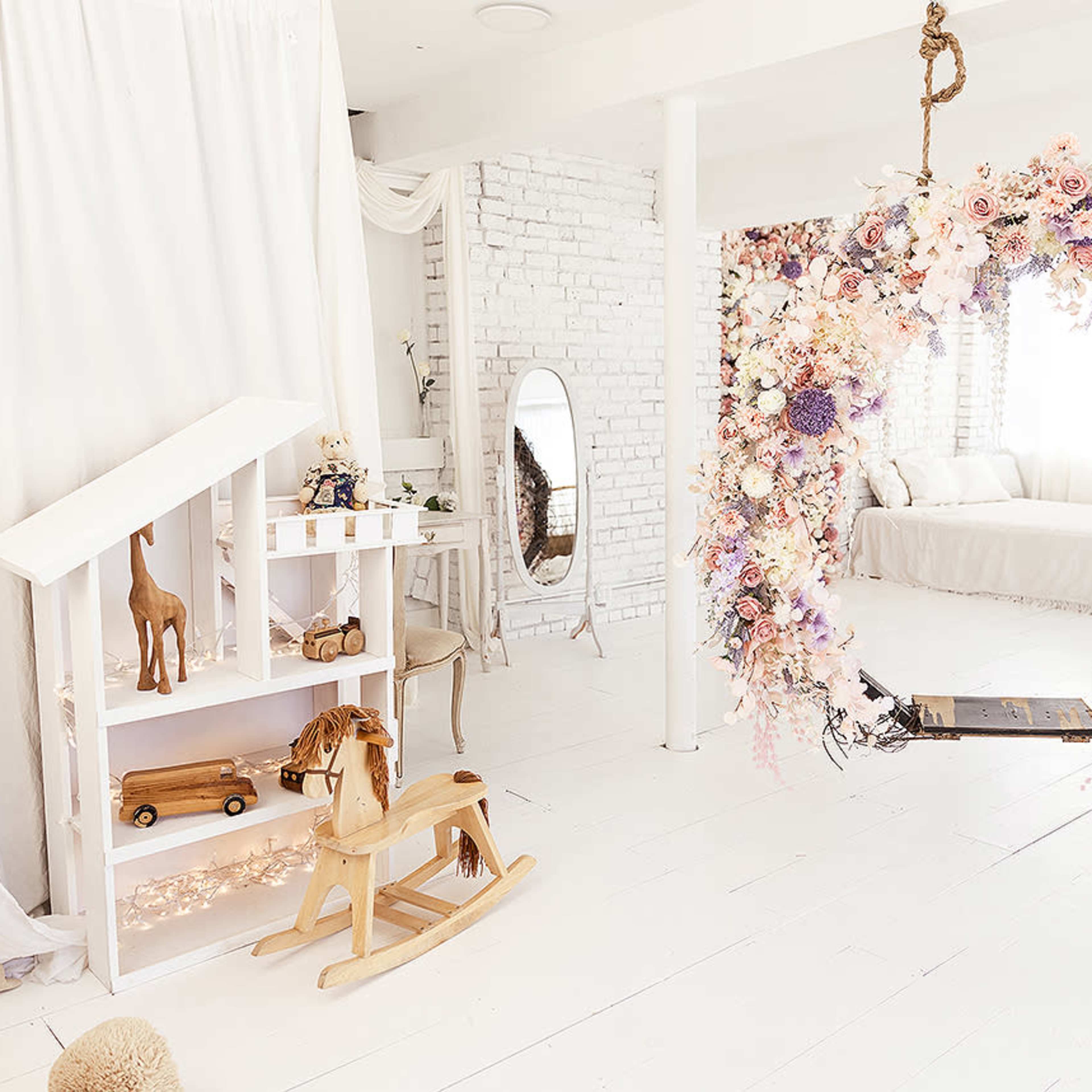 A bright, airy room with a floral swing, a white wooden playhouse, and a rocking horse.