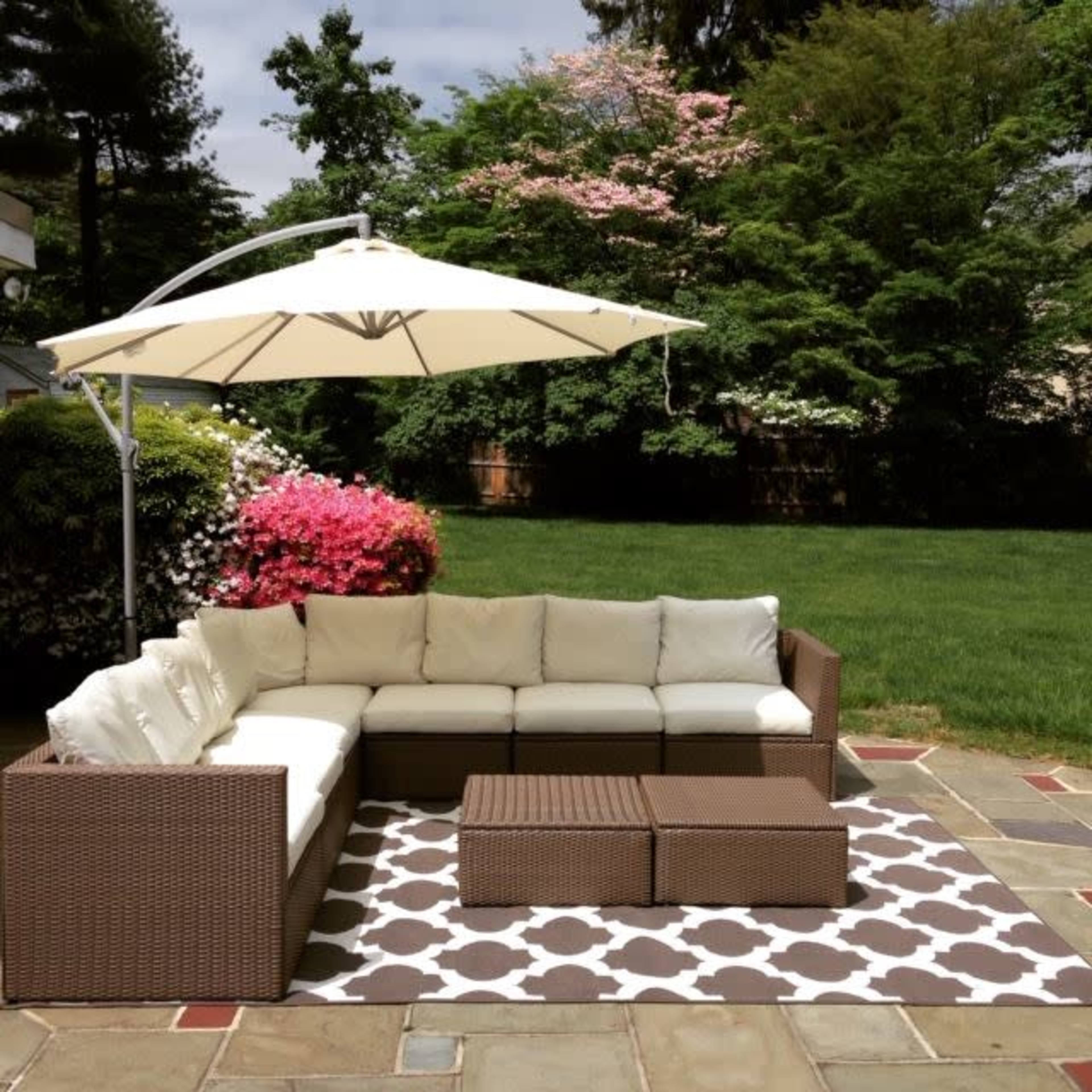 A sectional outdoor seating arrangement with white cushions is positioned on a patterned rug under a large umbrella in a garden with green grass and flowering bushes.