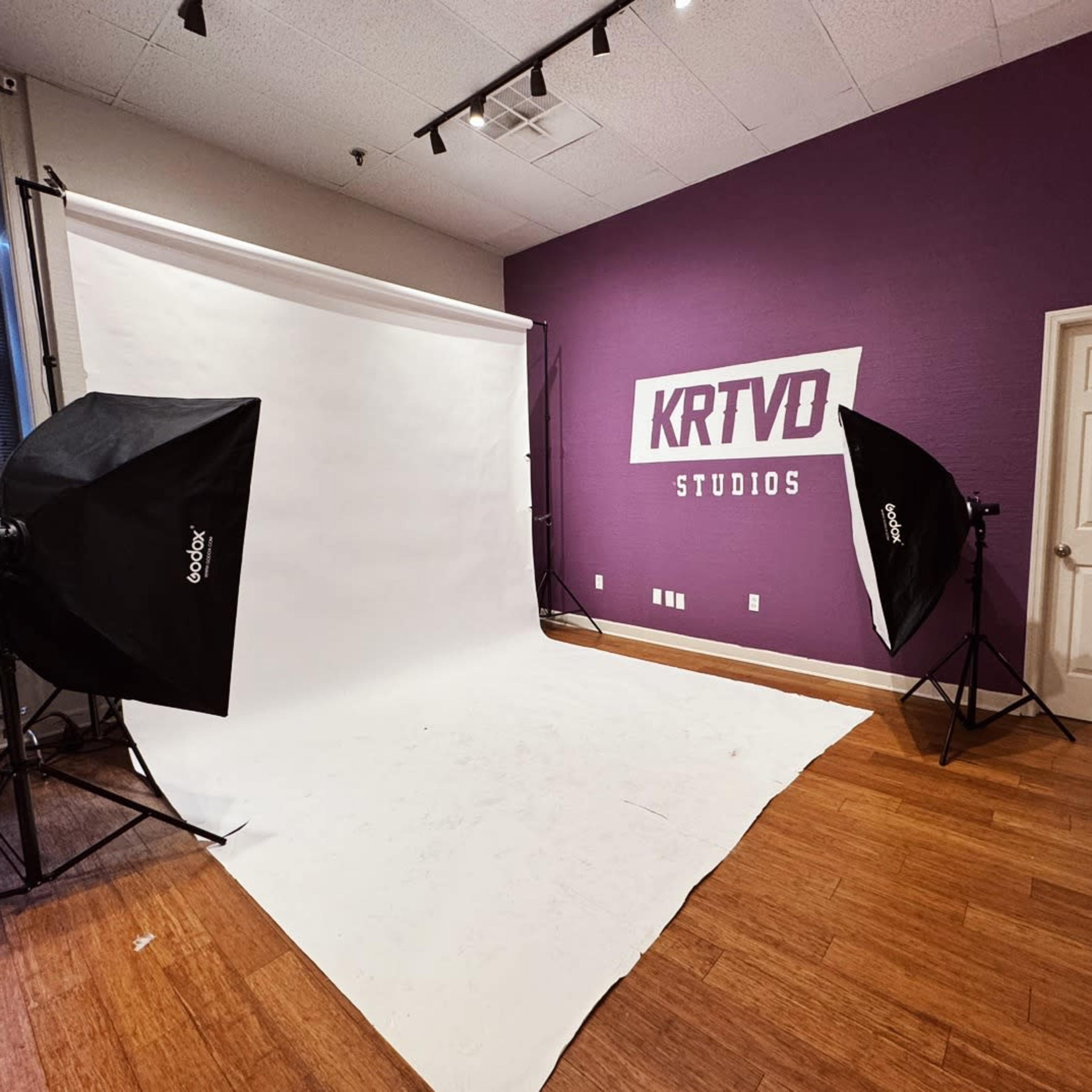 A photography studio featuring a white backdrop and two softbox lights set up against a purple wall with the text "KRTVD STUDIOS."