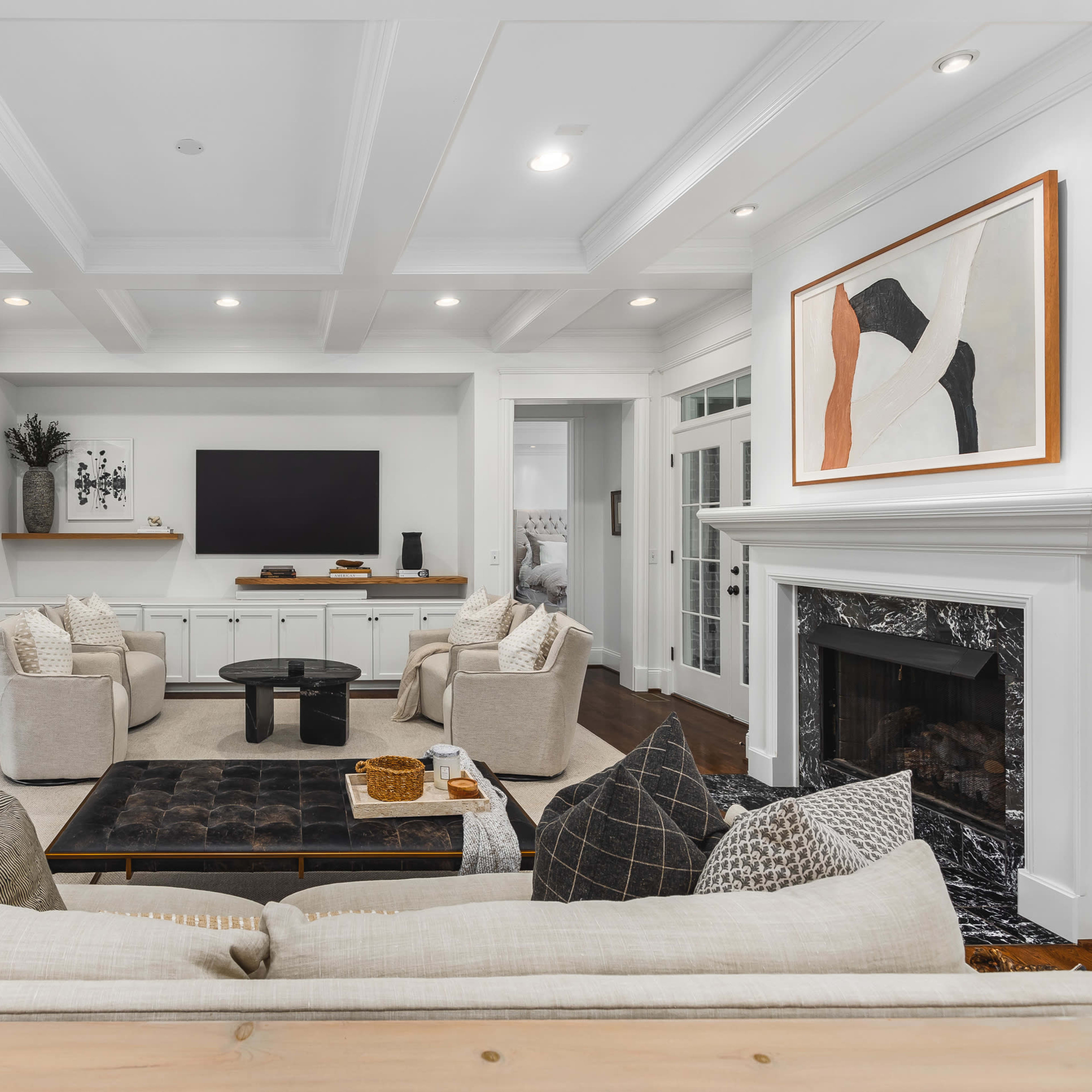 A modern living room features a central coffee table surrounded by plush armchairs and a fireplace, with a television mounted on the wall and artwork displayed nearby.