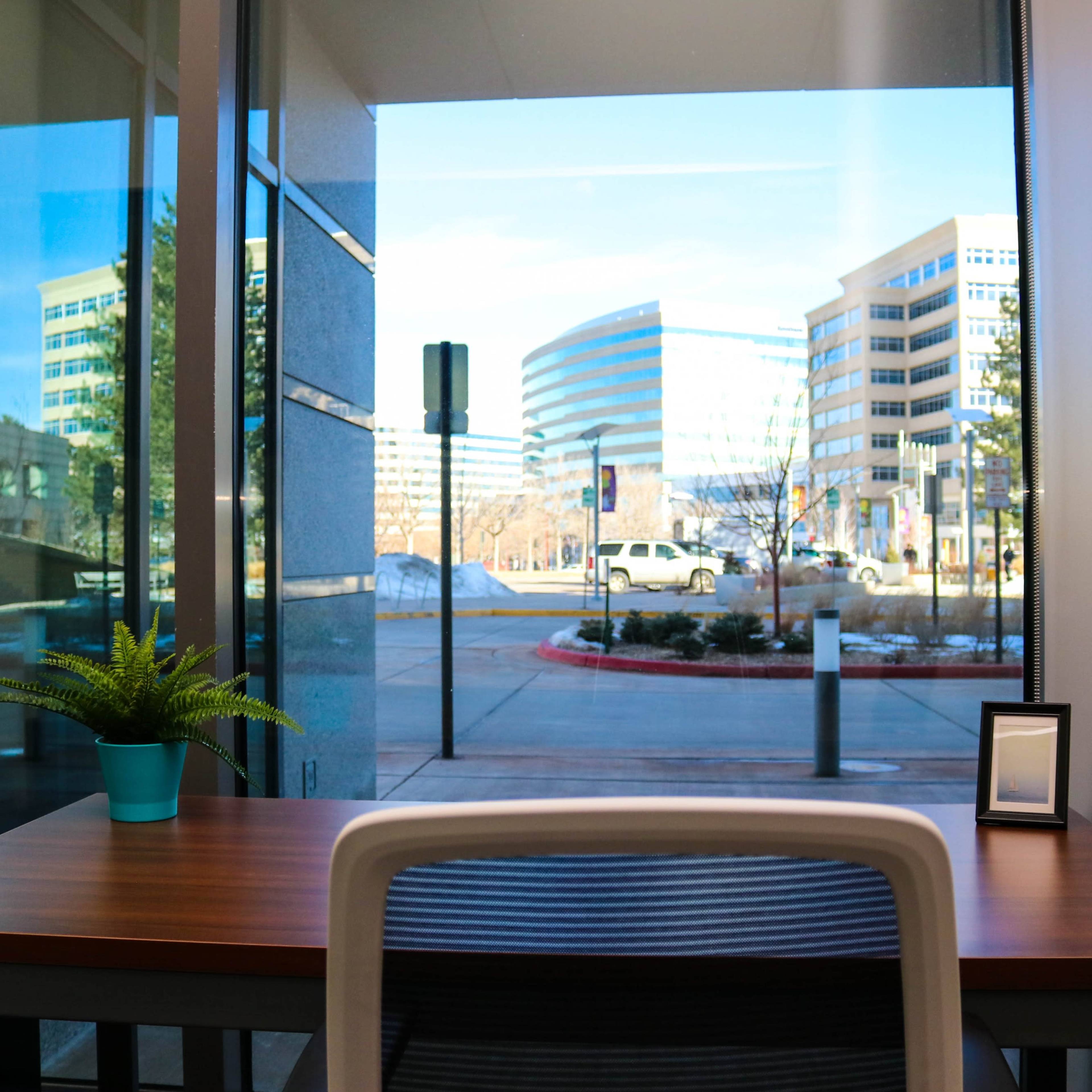 A modern workspace features a wooden table with a potted plant and two picture frames, overlooking a city street through large glass windows.
