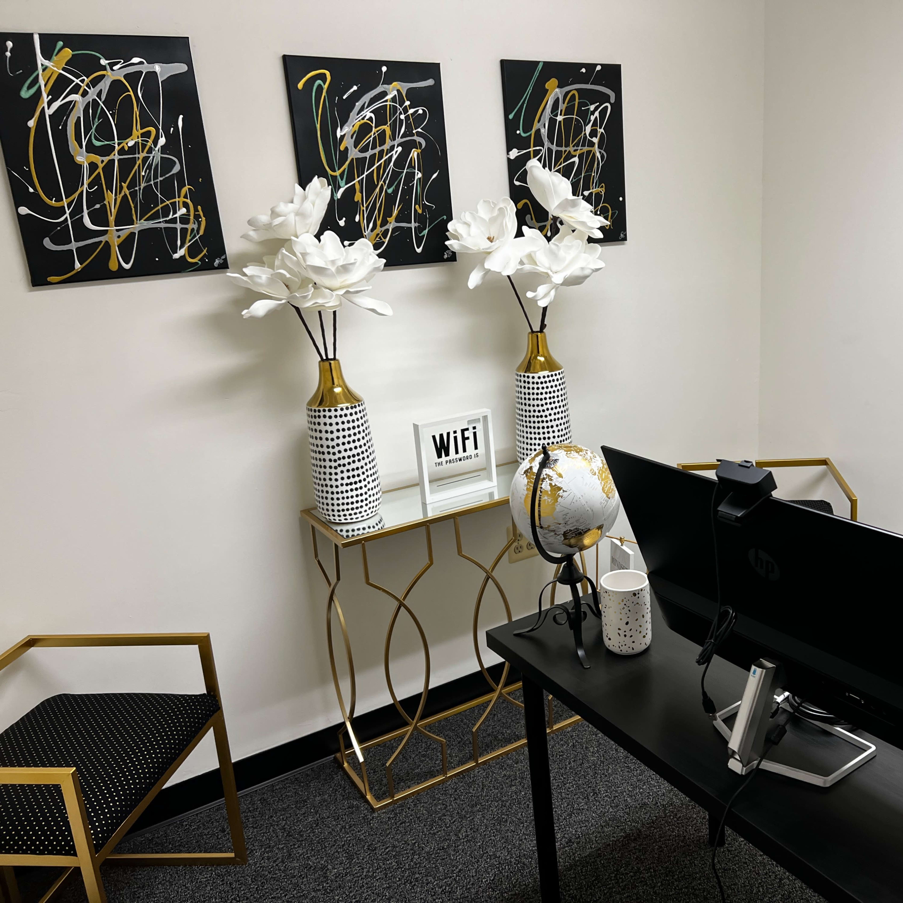 A modern office space featuring a black desk with a computer monitor, gold accents, and decorative elements including two white flower arrangements and framed abstract art on the walls.