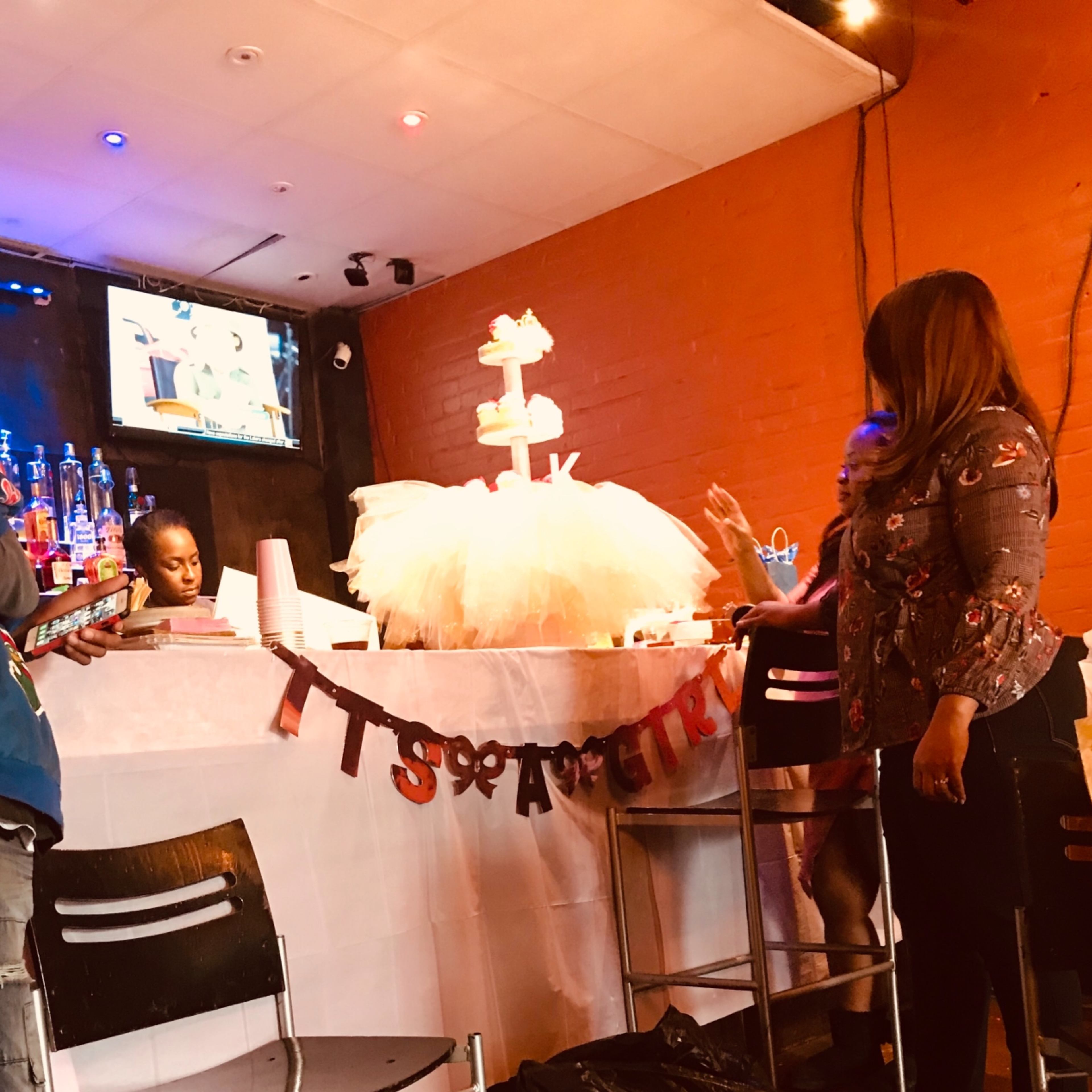 The scene shows a bar area with a decorated cake on the counter, a "It's a Girl" banner, and two people interacting nearby.
