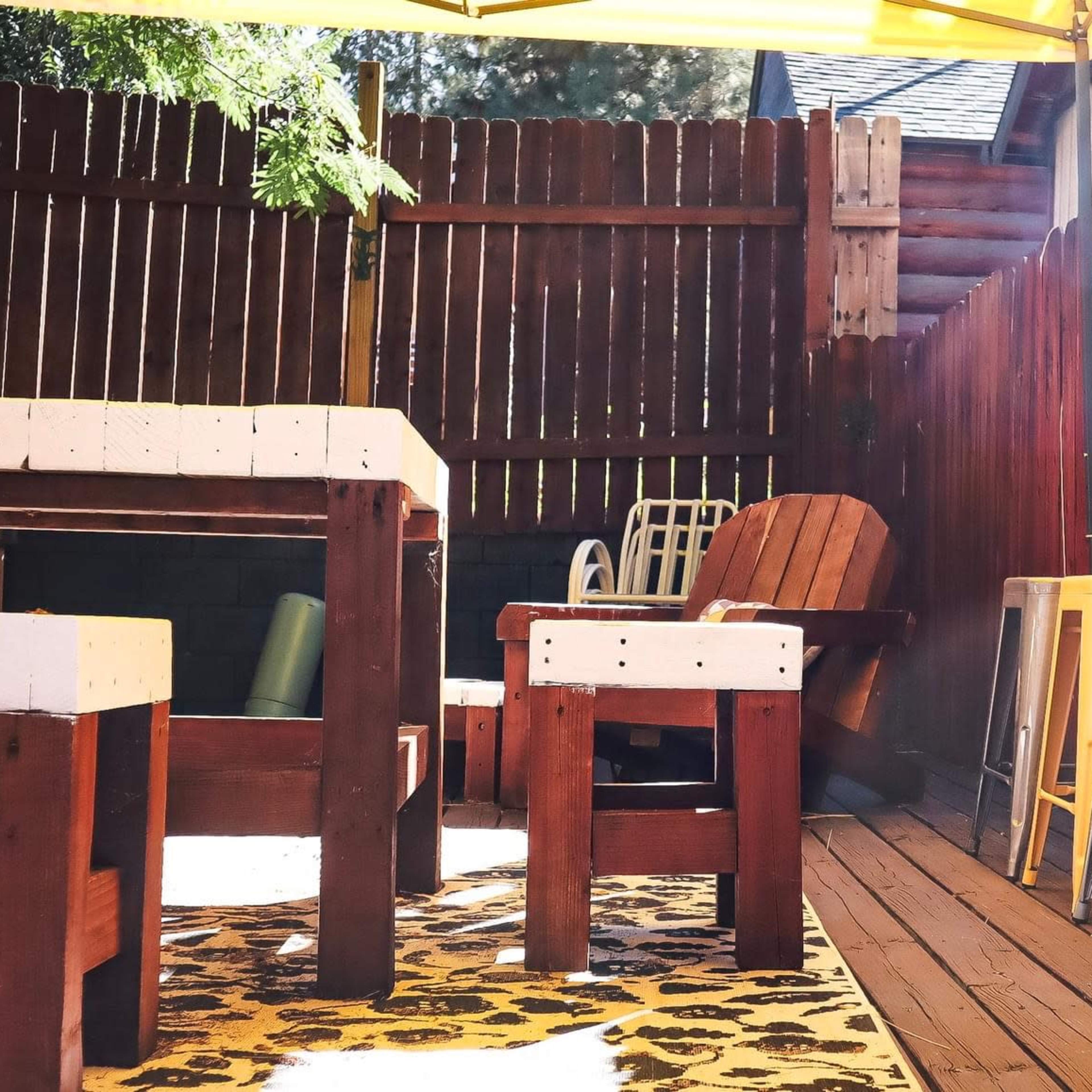An outdoor seating area featuring wooden furniture and bar stools under a yellow canopy, surrounded by a wooden fence.