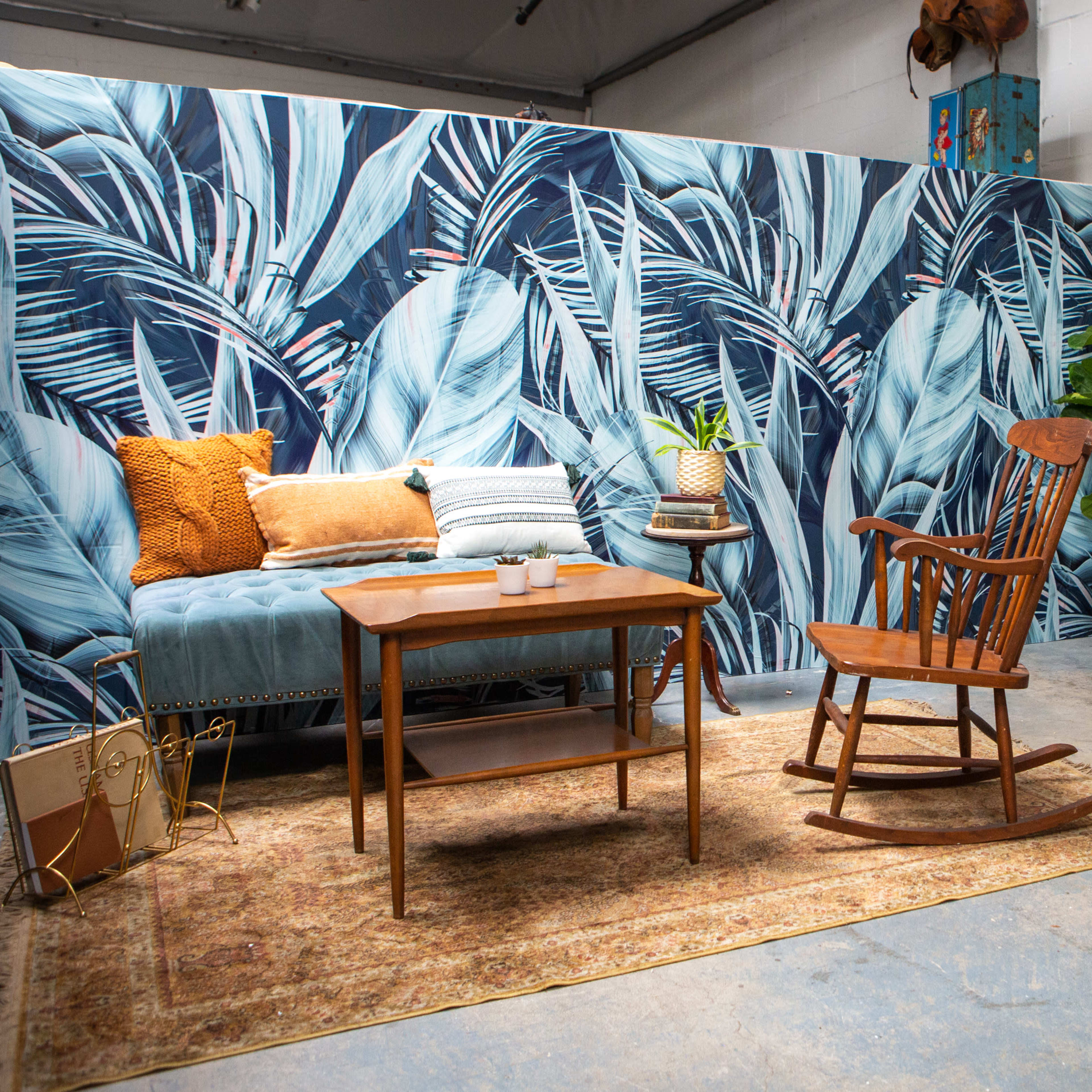 A cozy interior space featuring a wooden rocking chair, a small coffee table, and a writing desk against a backdrop of vibrant palm leaf wallpaper.