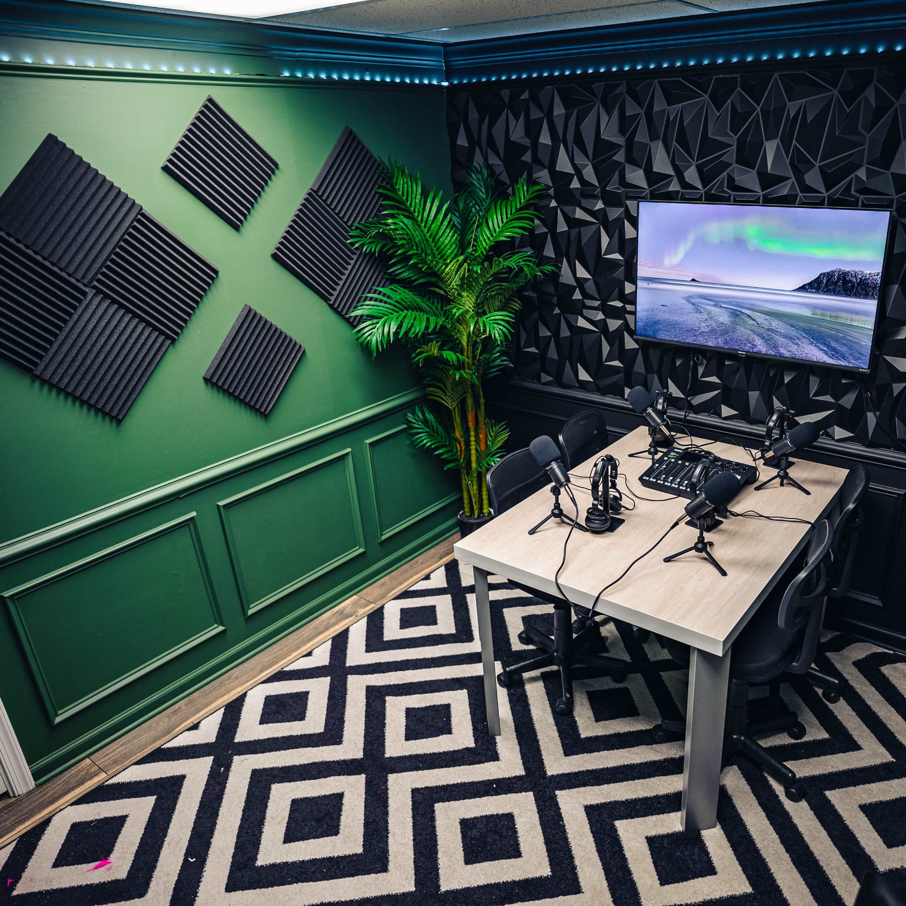 A modern recording studio with two microphones positioned on a table, a large monitor displaying a scenic image, and soundproofing panels on the walls.