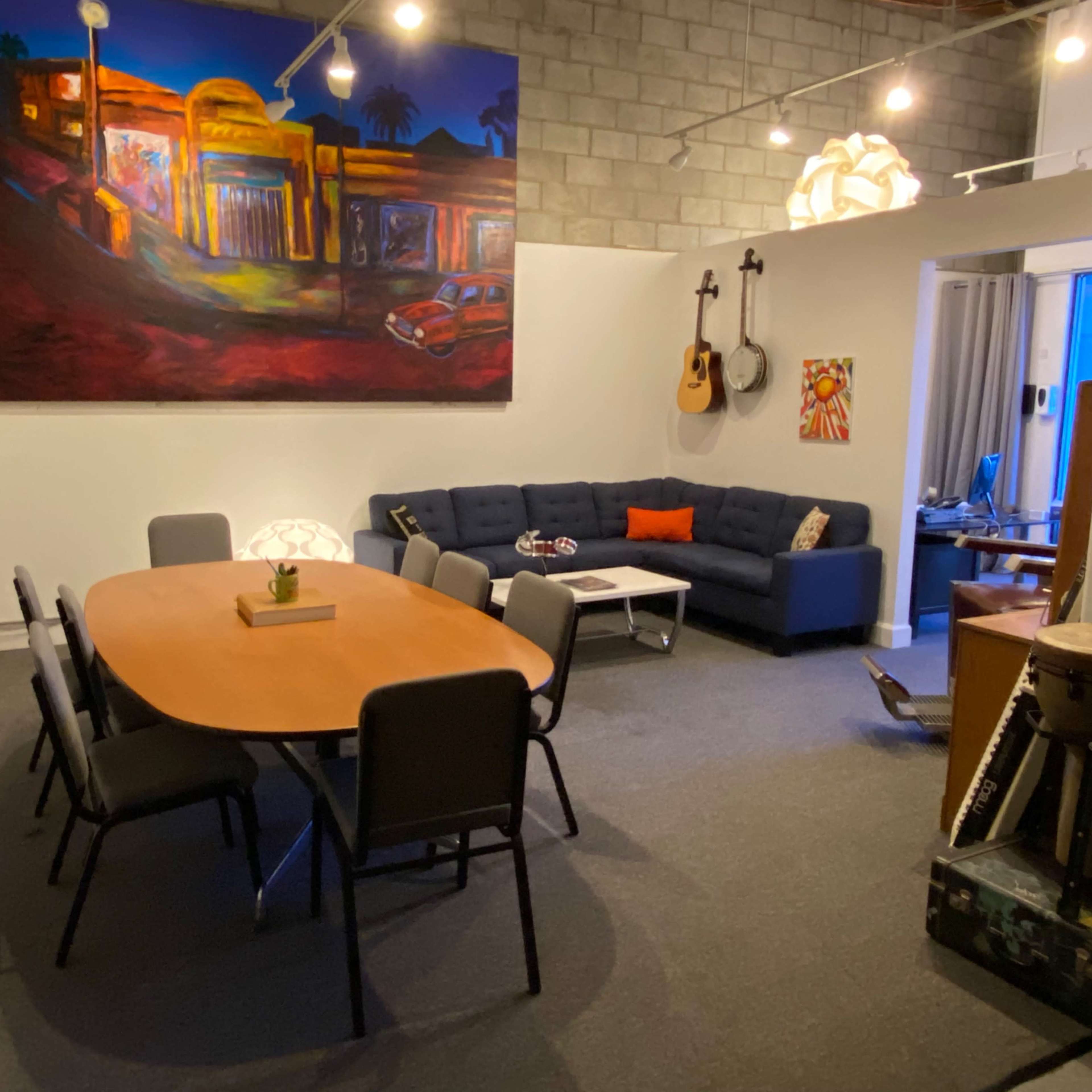 A modern meeting space featuring a large oval table surrounded by chairs, a blue sectional sofa, an artwork on the wall, and various musical instruments.