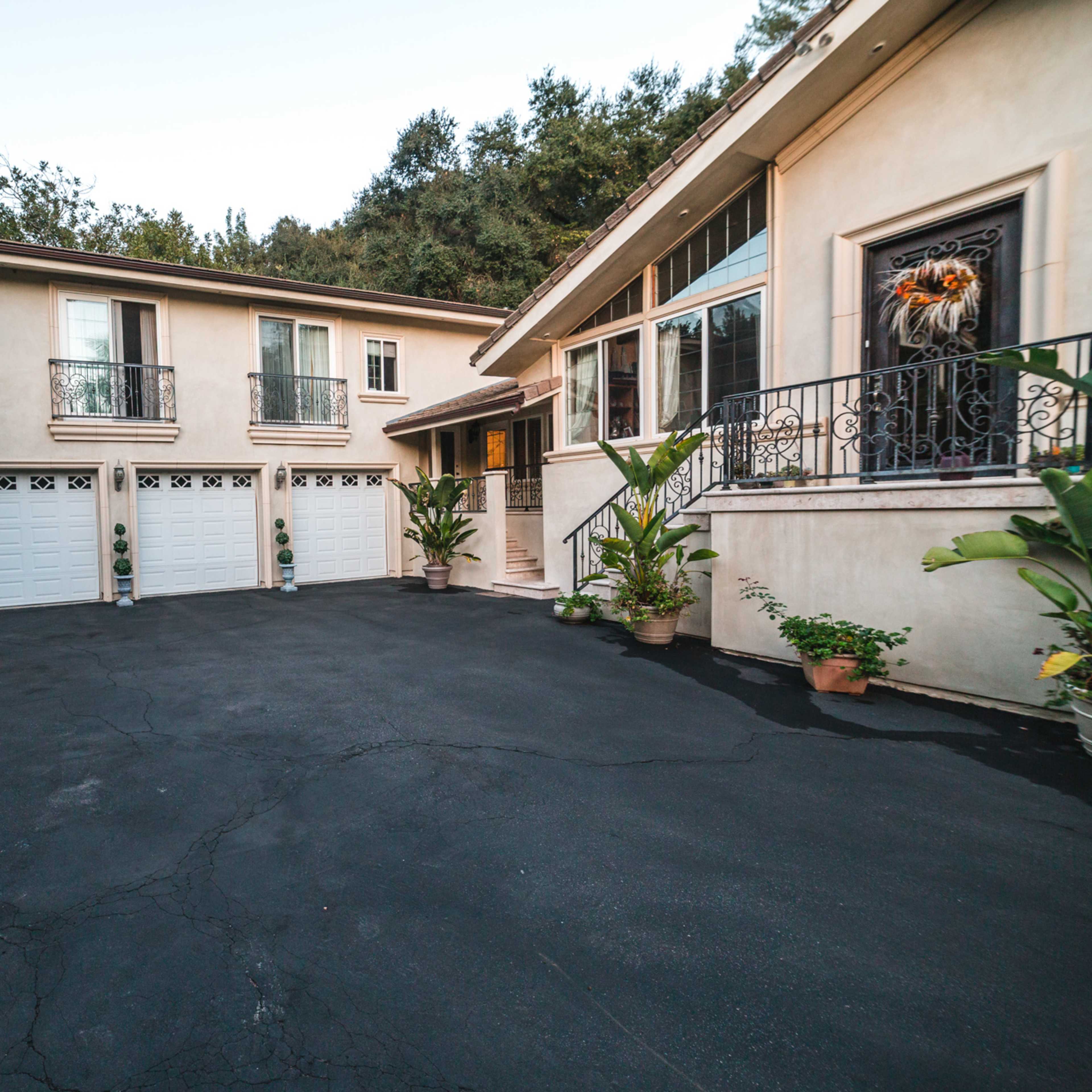 A two-story residential building with a paved driveway leading to a series of garage doors, surrounded by potted plants and outdoor seating.
