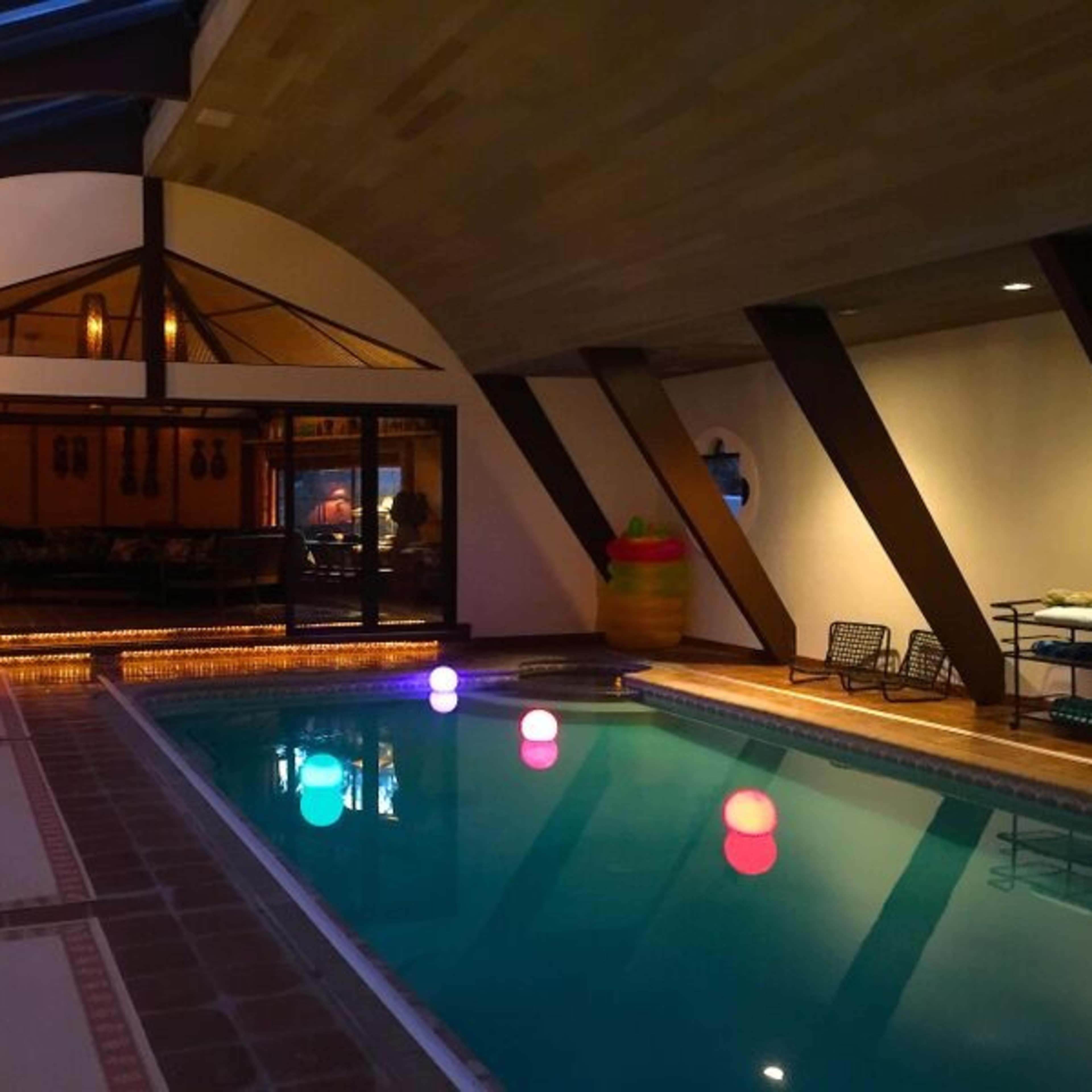 An indoor pool area features colorful floating lights and a large window overlooking a dining space.