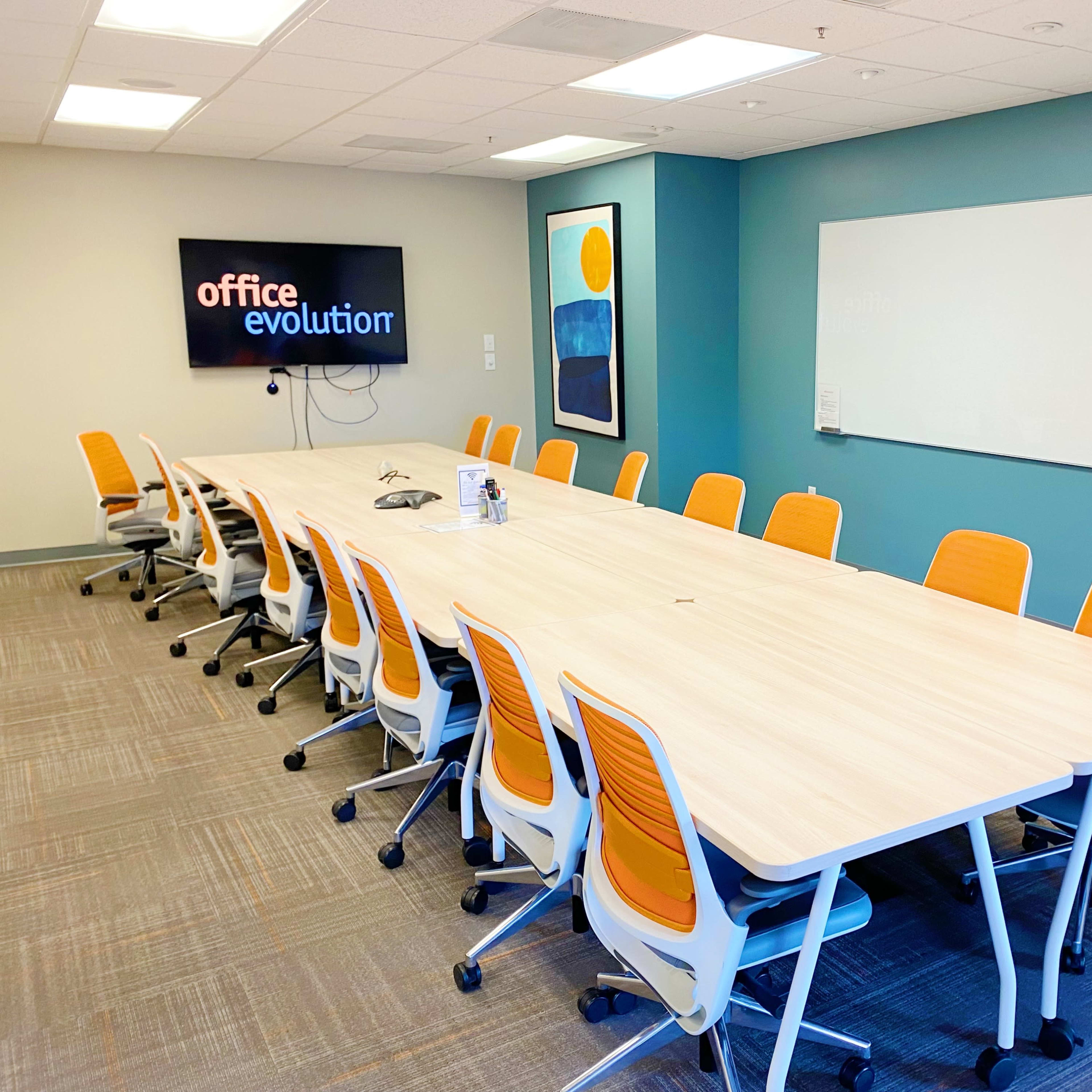 A large conference room features a long wooden table surrounded by orange and white chairs, with a screen displaying "office evolution" on the wall and a whiteboard nearby.