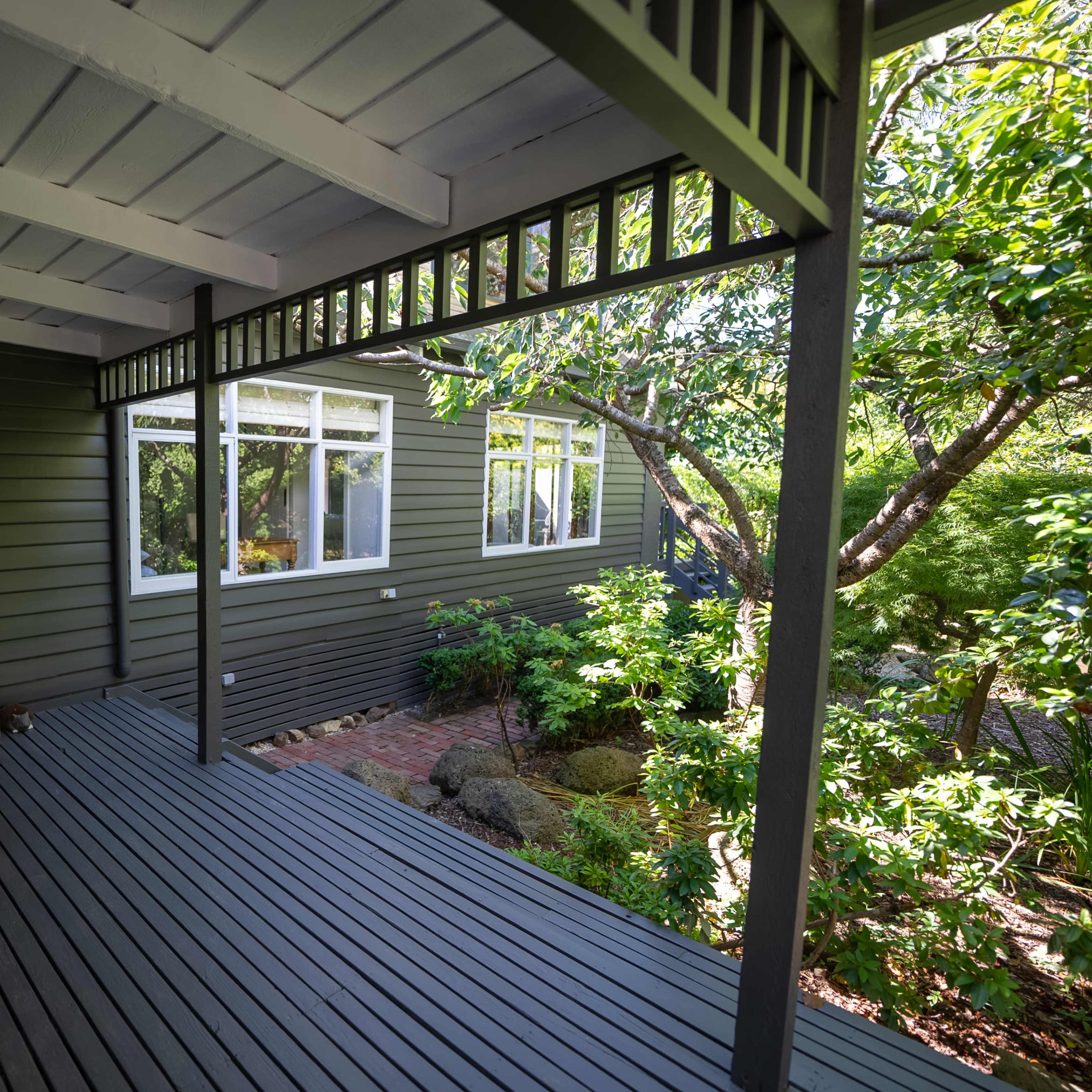 A shaded porch with wooden flooring leads to a house featuring green siding and large windows surrounded by lush foliage.
