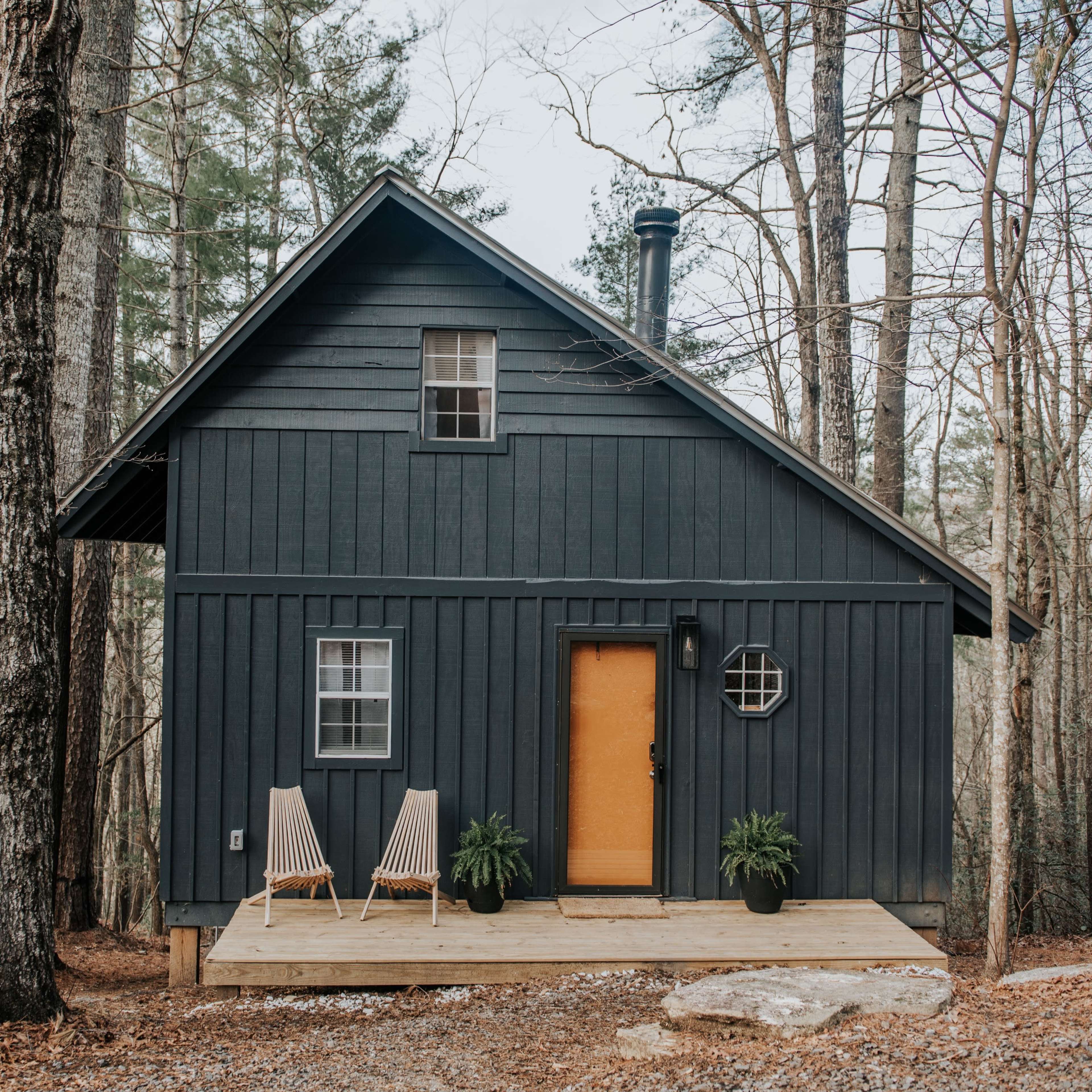 A dark wooden cabin with a yellow door and two chairs sits on a wooden porch surrounded by trees.