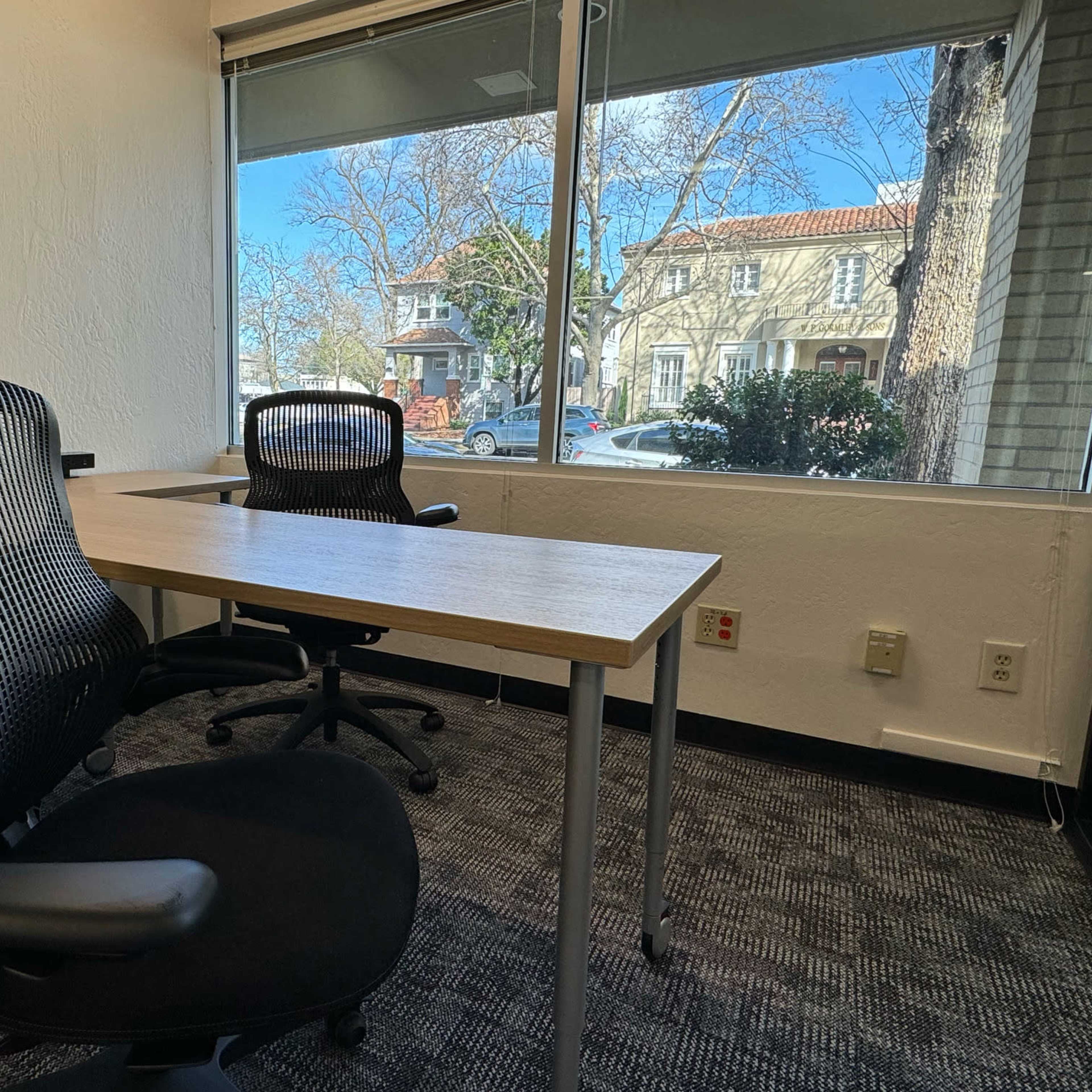 A modern office workspace features a wooden table and two mesh chairs, with a view of a tree-lined street outside.