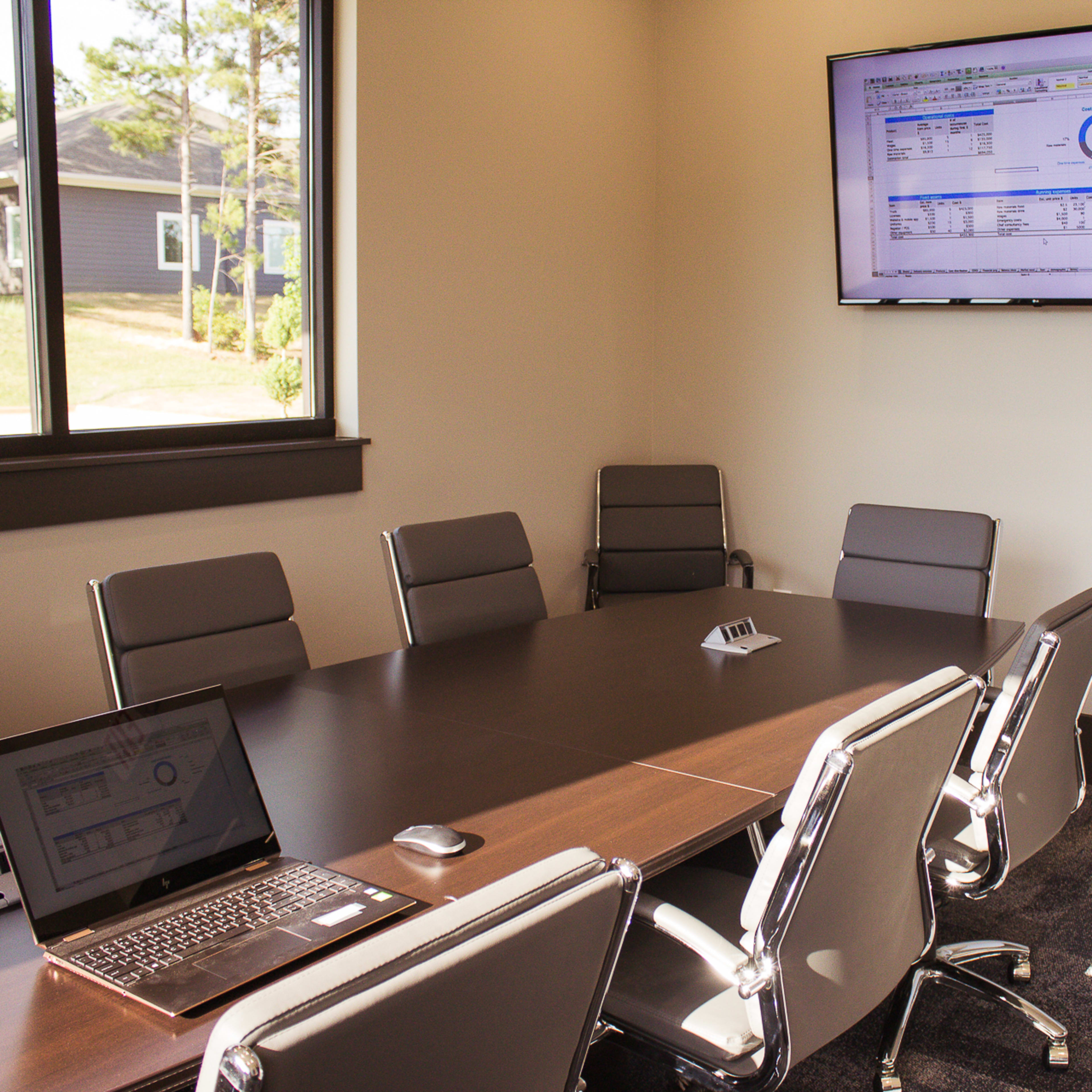 A modern conference room features a large table, eight chairs, a laptop on the table, a presentation screen on the wall, and a whiteboard.