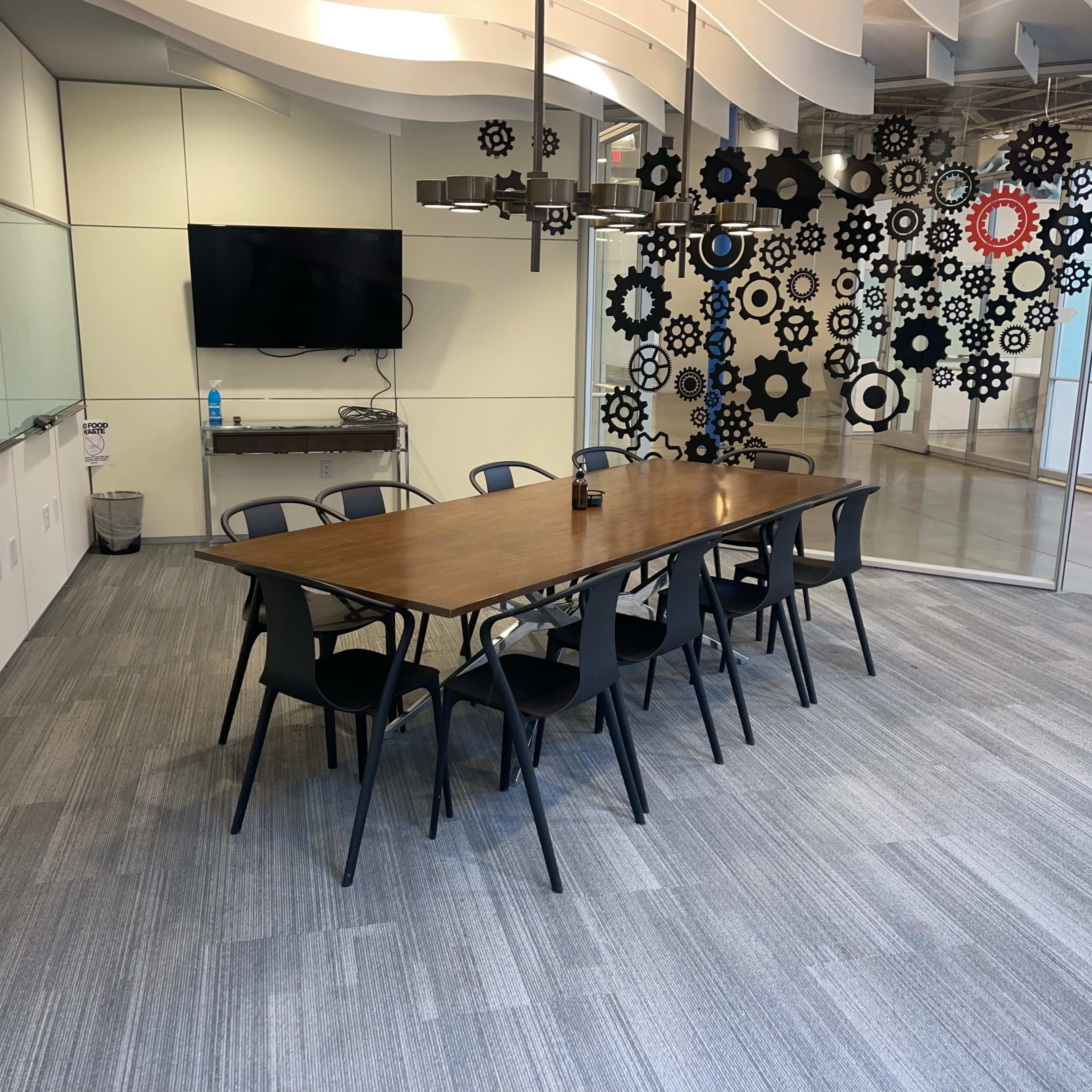 A meeting room features a long wooden table surrounded by black chairs, with a television on one wall and decorative gear shapes on another.