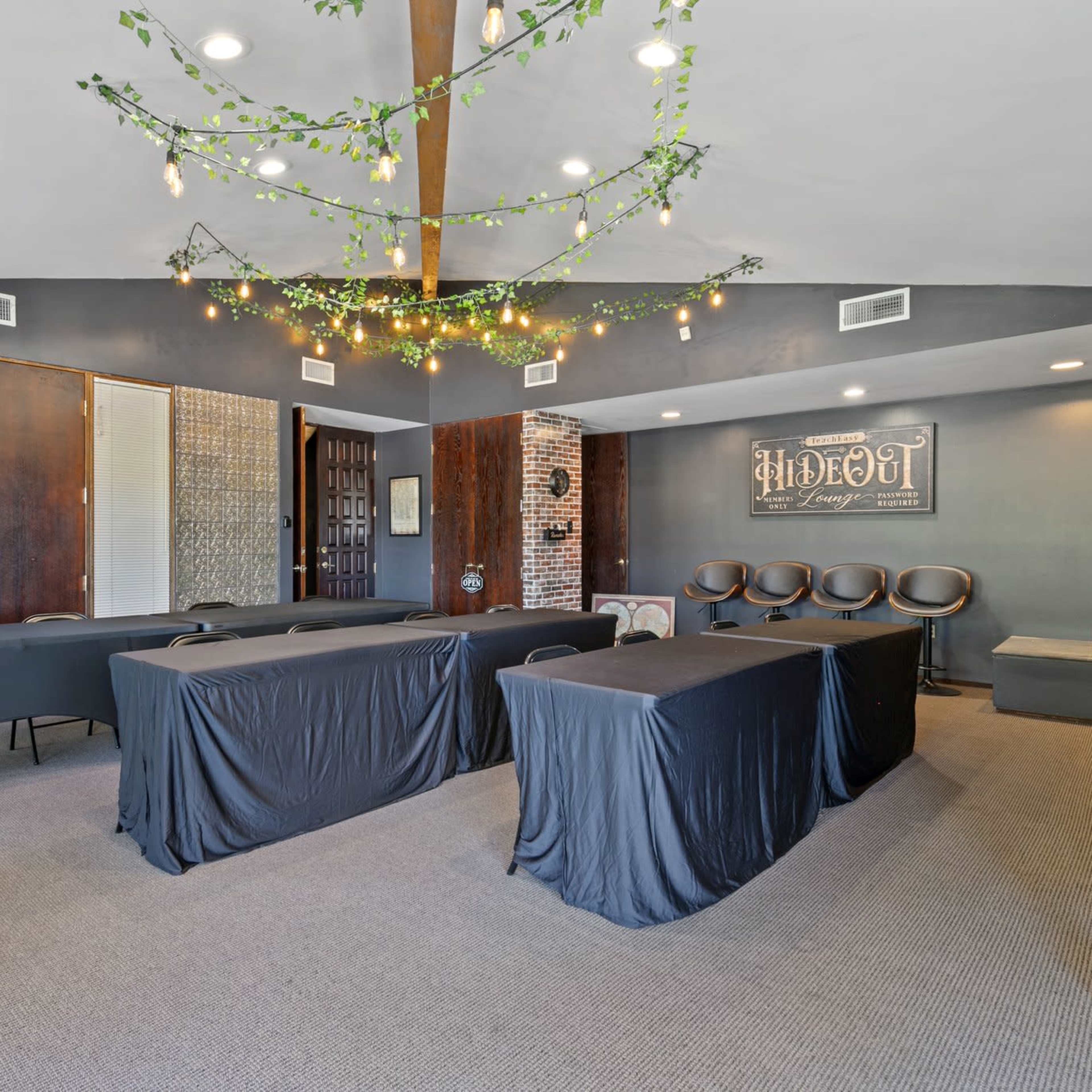 A conference room features several tables covered with black linens, surrounded by chairs, and decorated with hanging greenery and lights.