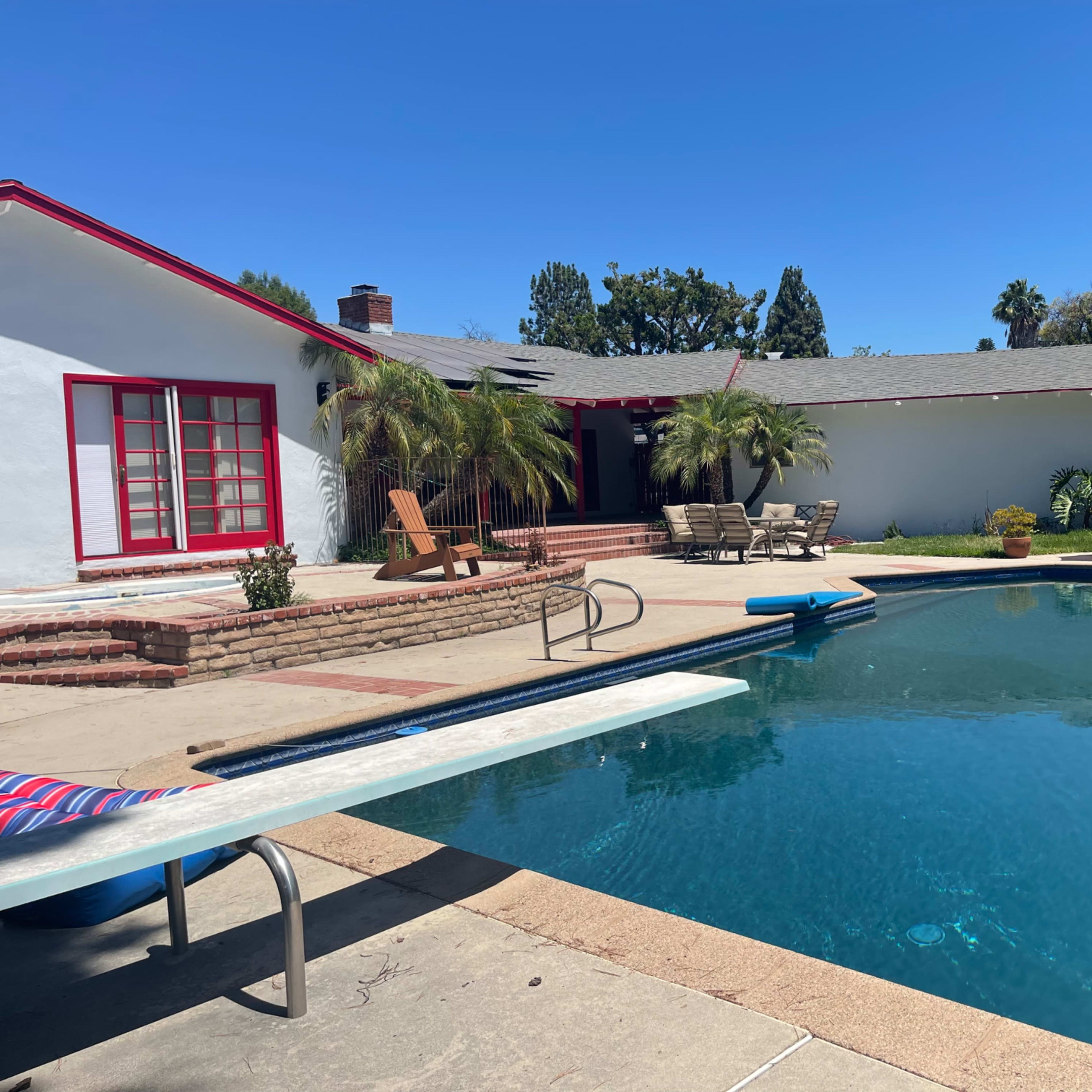 A backyard pool area with a diving board, lounge chairs, an umbrella, and a house in the background.
