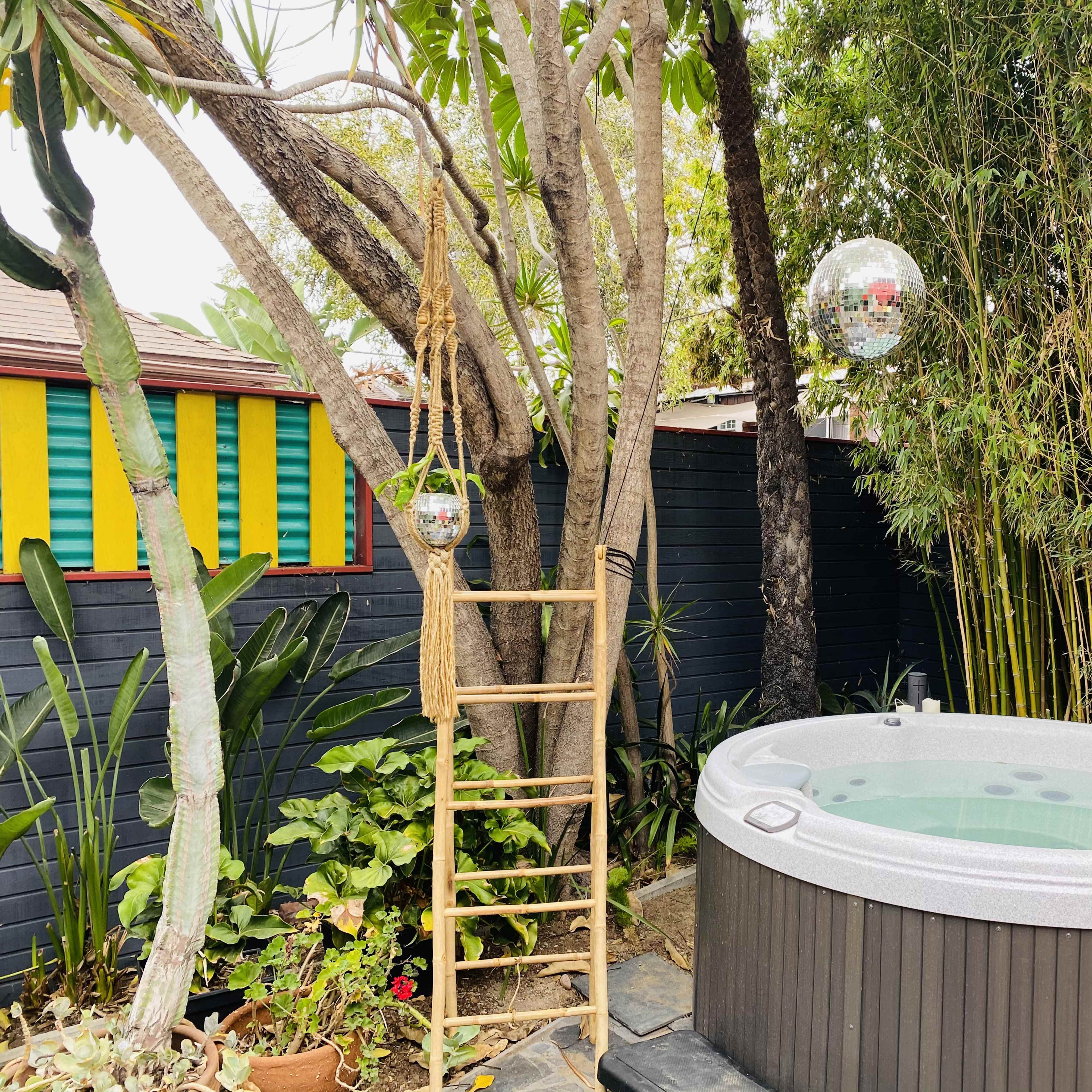 A hot tub is situated next to a bamboo grove, with a wooden ladder leaning against a tree and disco balls hanging above.