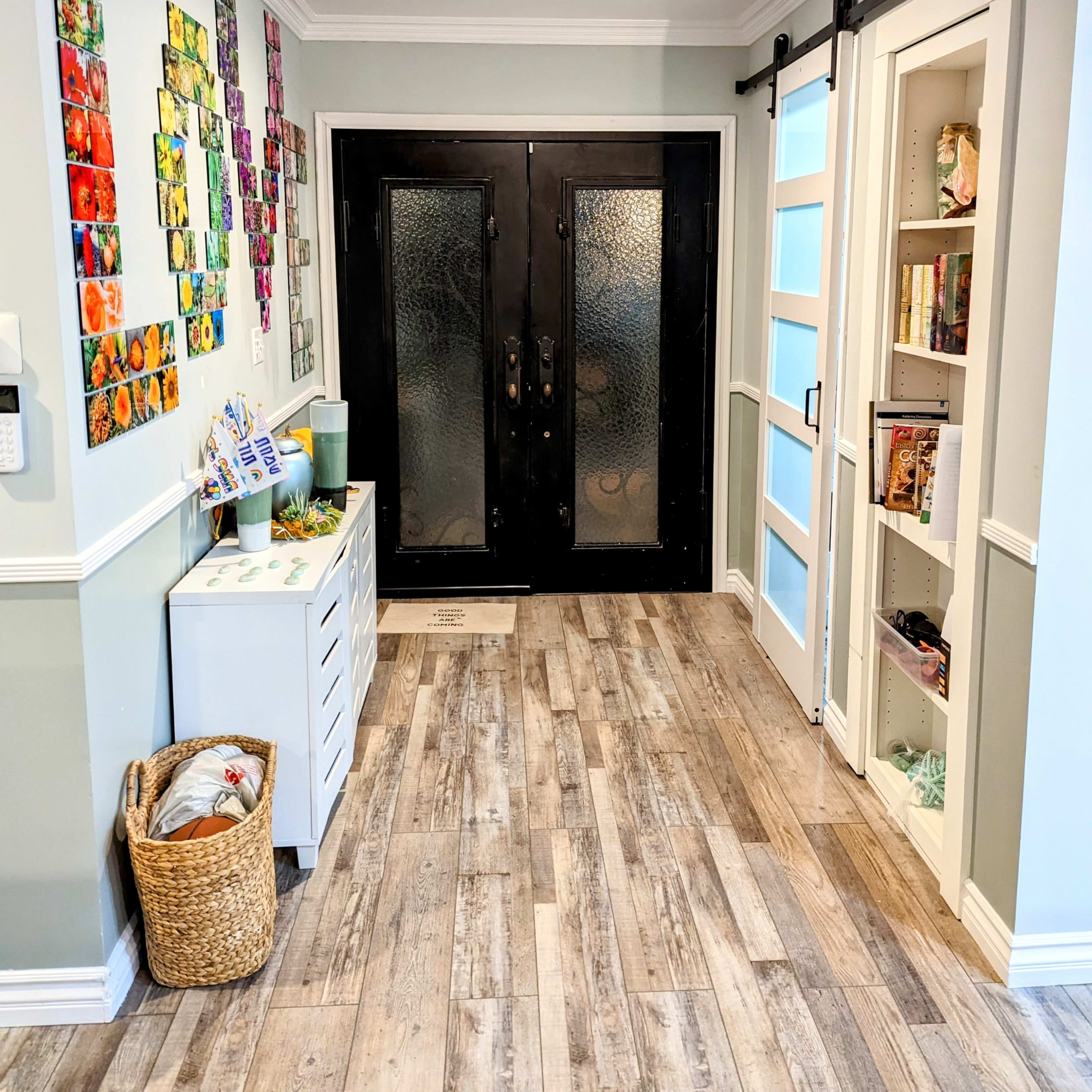 A well-lit entryway with black double doors, a decorated wall of colorful artwork, and a small storage cabinet beside a shelf.