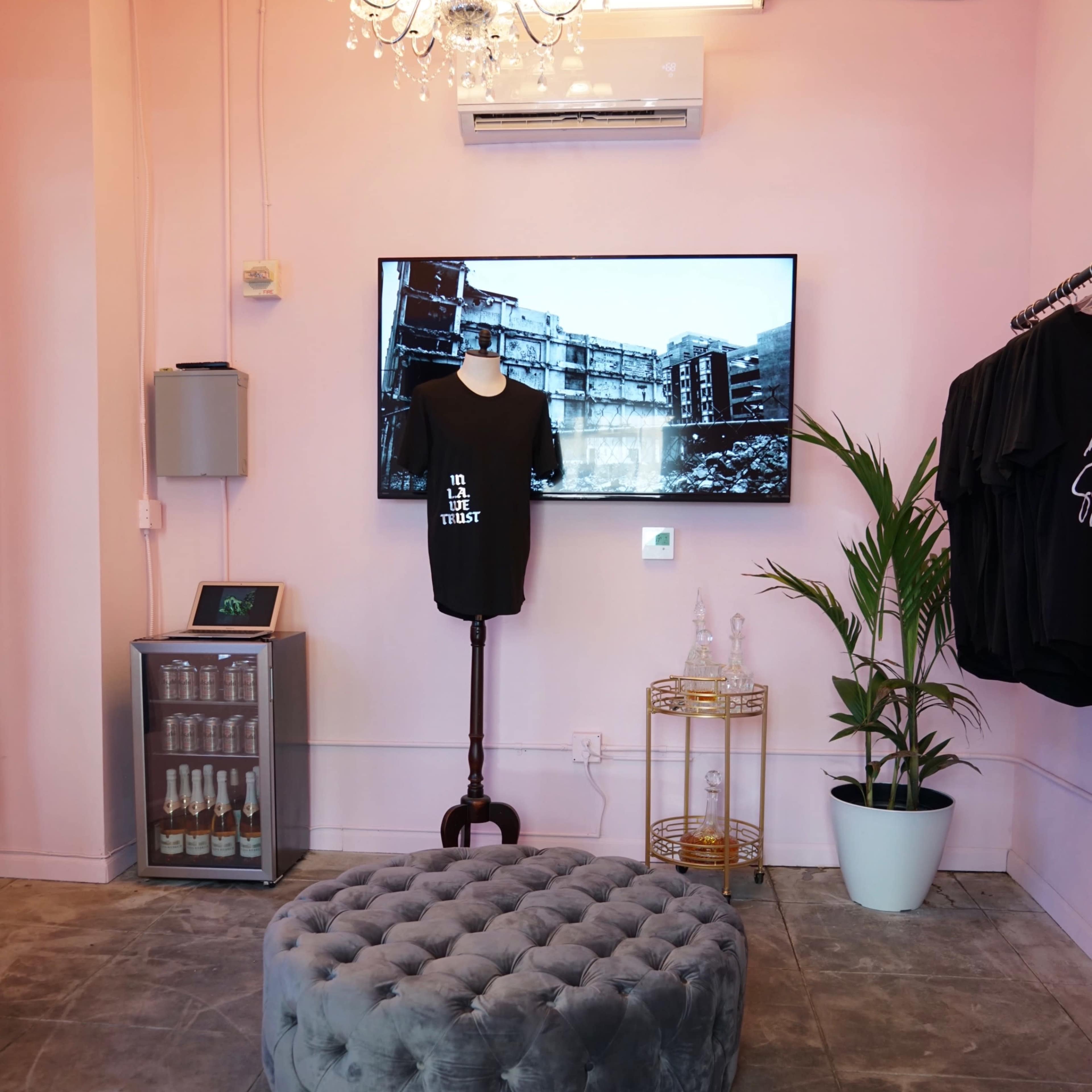 A pink-walled boutique featuring a tufted gray ottoman, a wall-mounted television, a plant in a white pot, and clothing displayed on hangers.