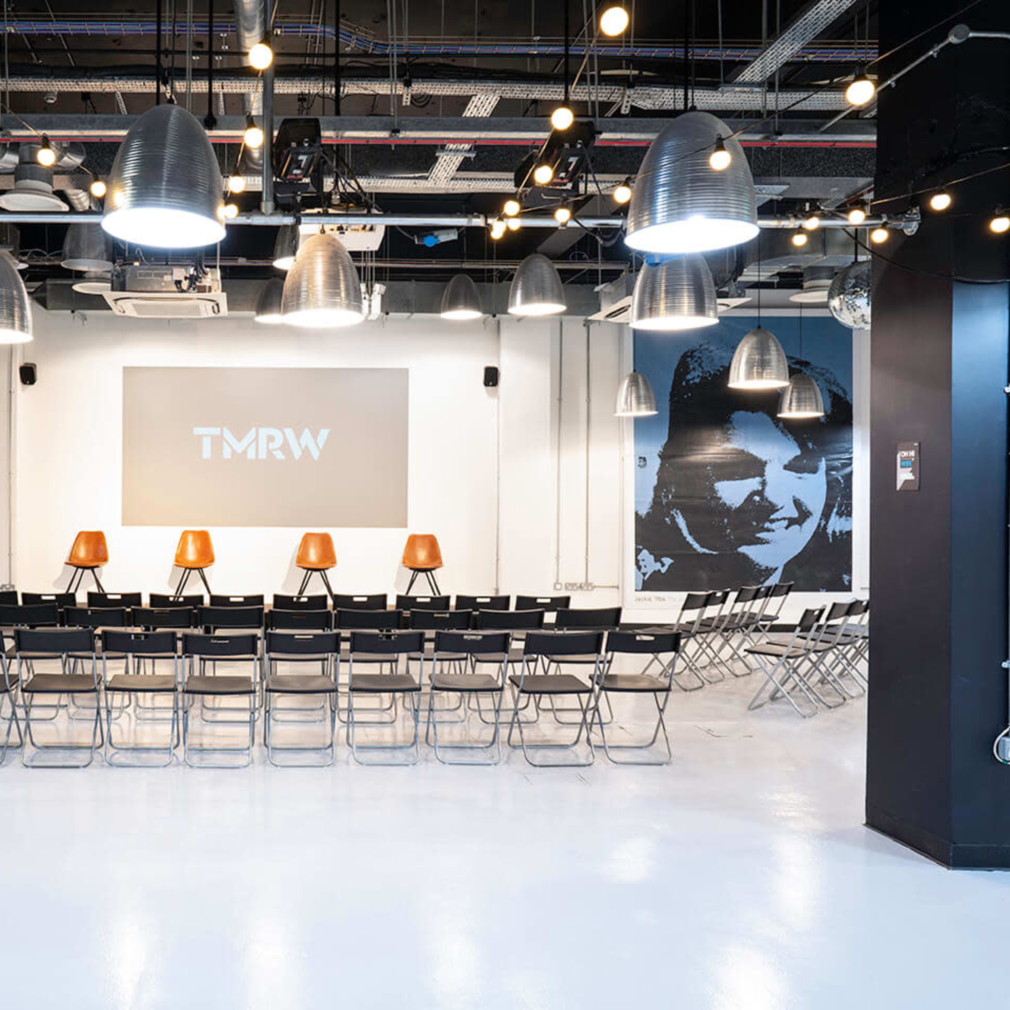 A modern event space with rows of black folding chairs facing a large screen, illuminated by pendant lights and featuring murals on the walls.