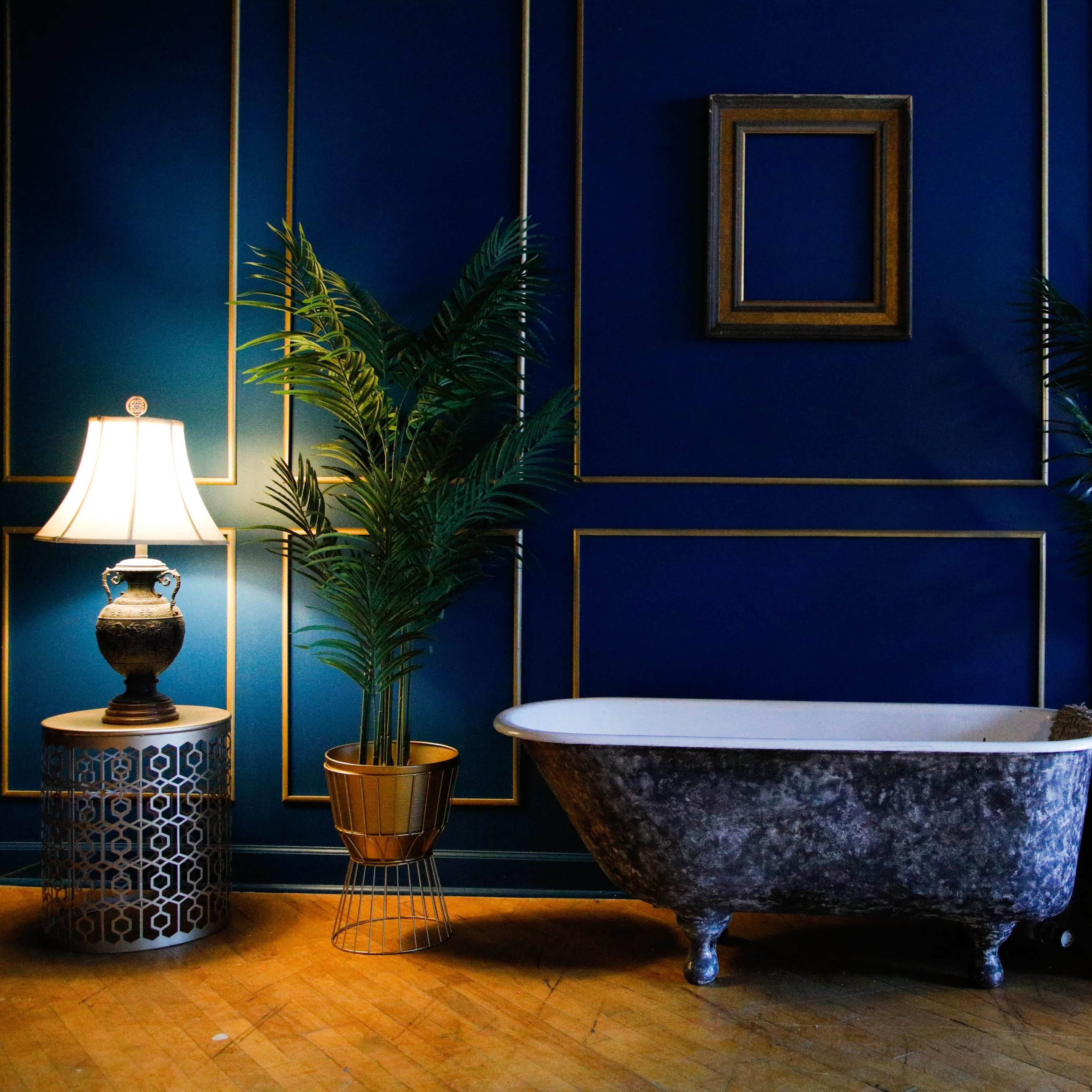 A vintage bathtub is placed against a dark blue wall adorned with gold trim, accompanied by potted plants, a lamp, and a striped chair.