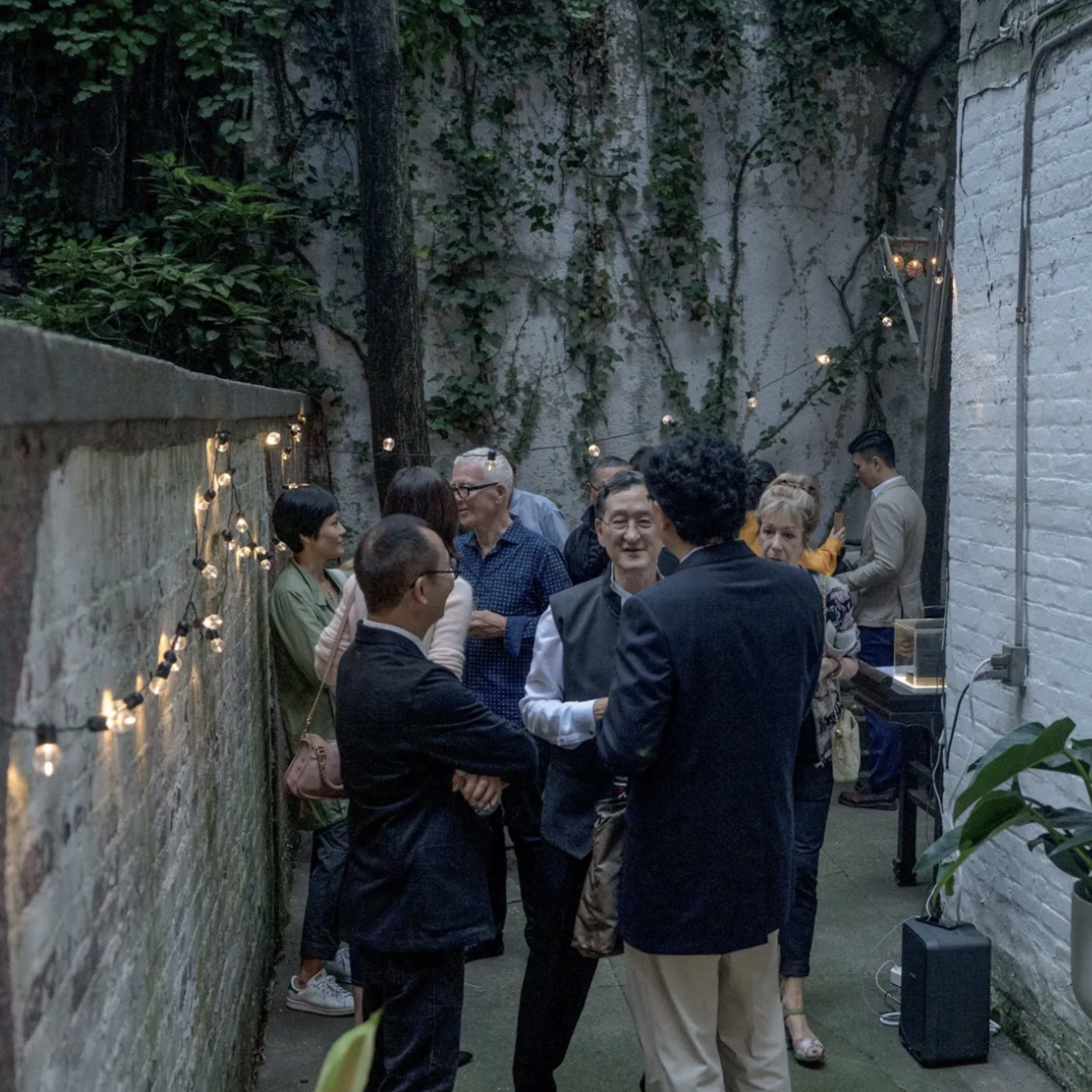 The scene shows a group of people engaging in conversation in a narrow outdoor space adorned with string lights and greenery.
