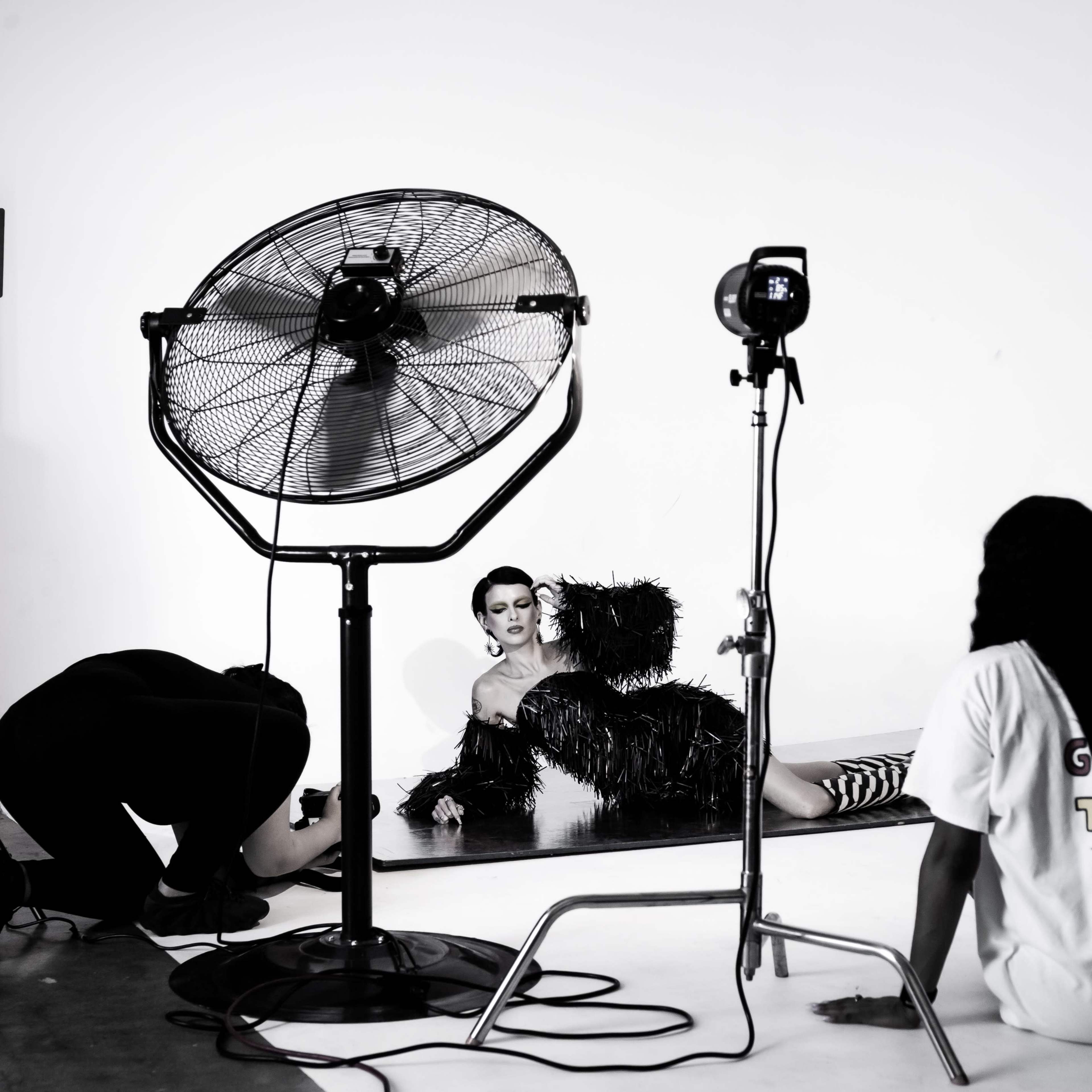 A model poses on a studio set while a large fan and lighting equipment are positioned nearby.