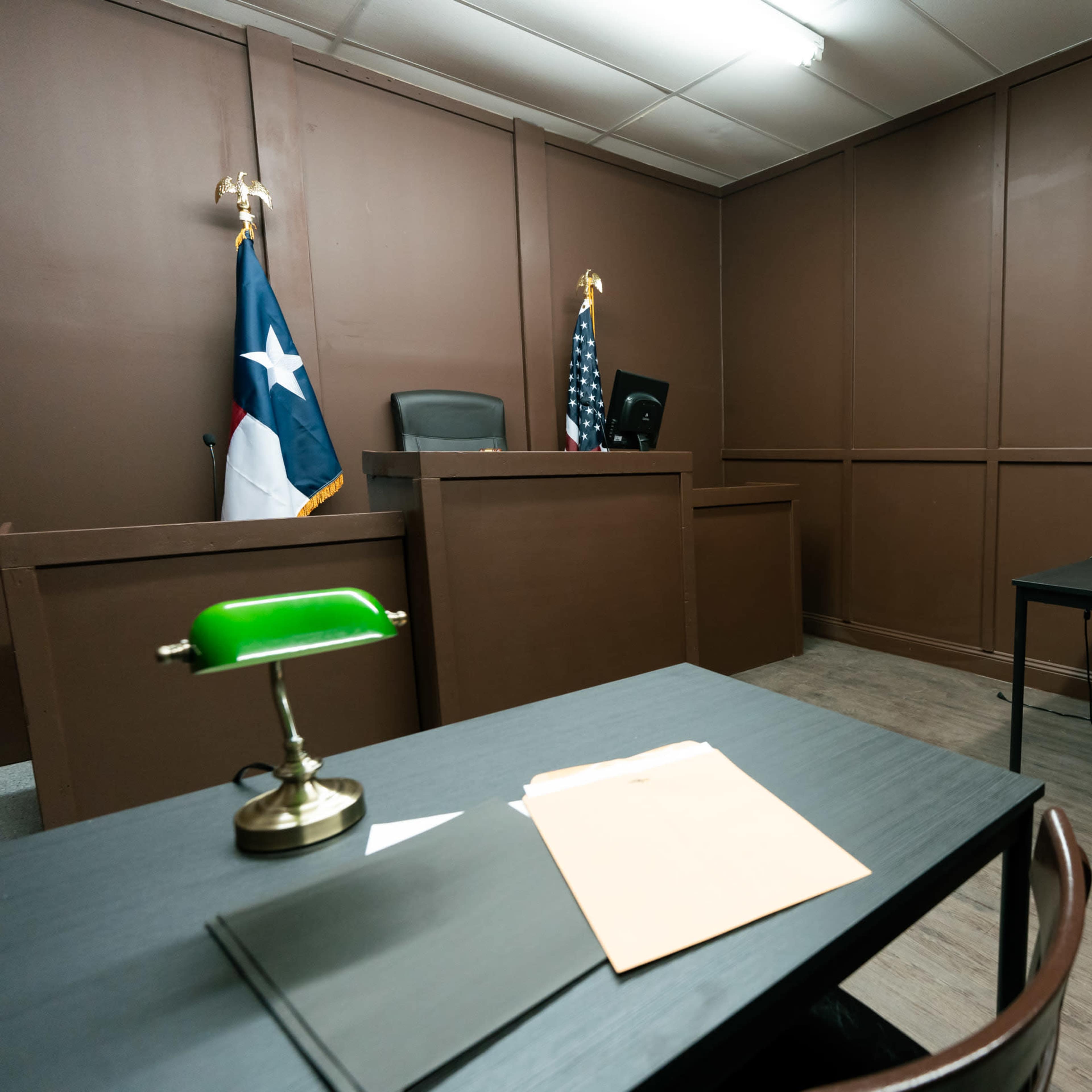 A small courtroom with a judge's bench, American and Texas flags, and two tables with chairs.
