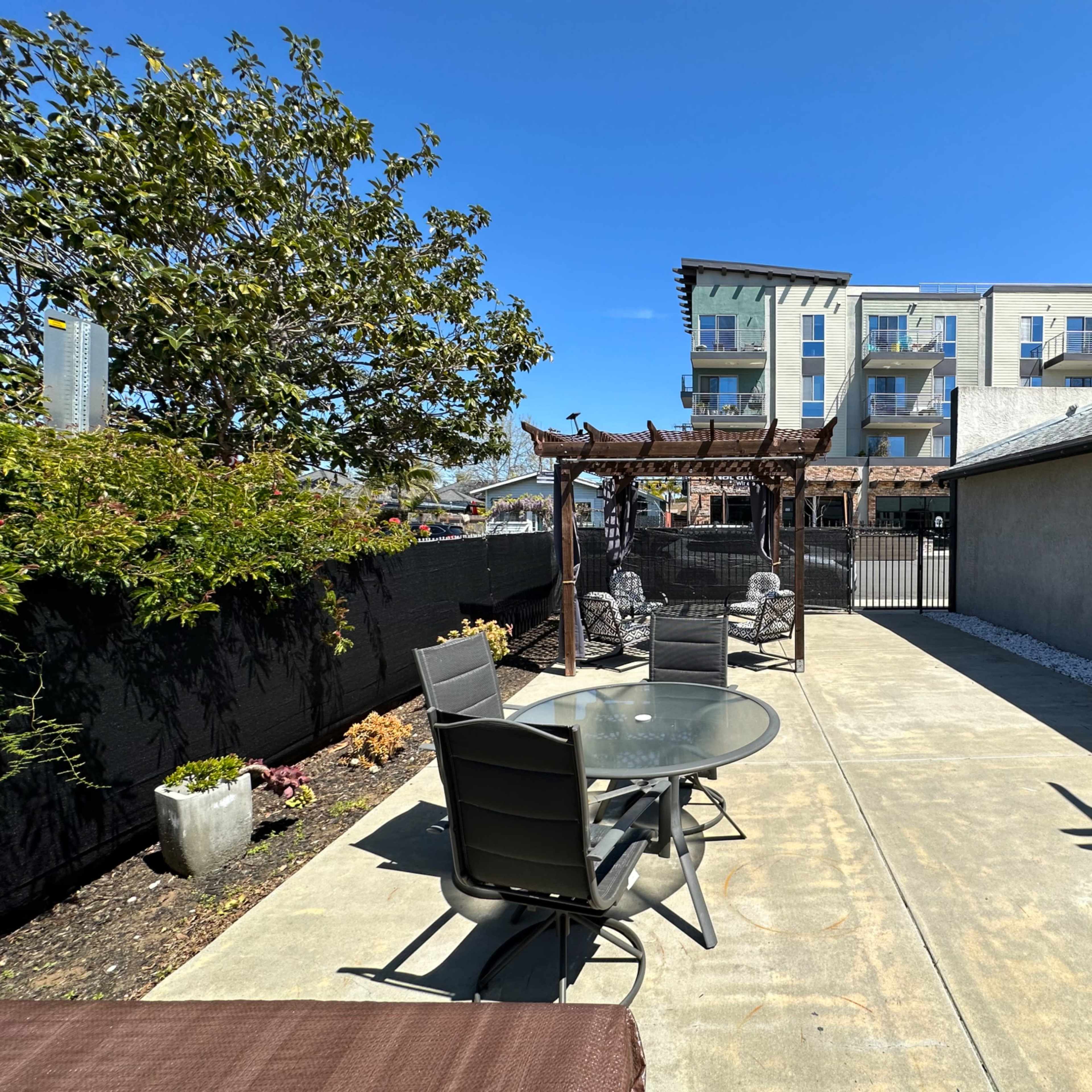 A concrete patio area with a glass table and chairs, a pergola, and a building in the background.
