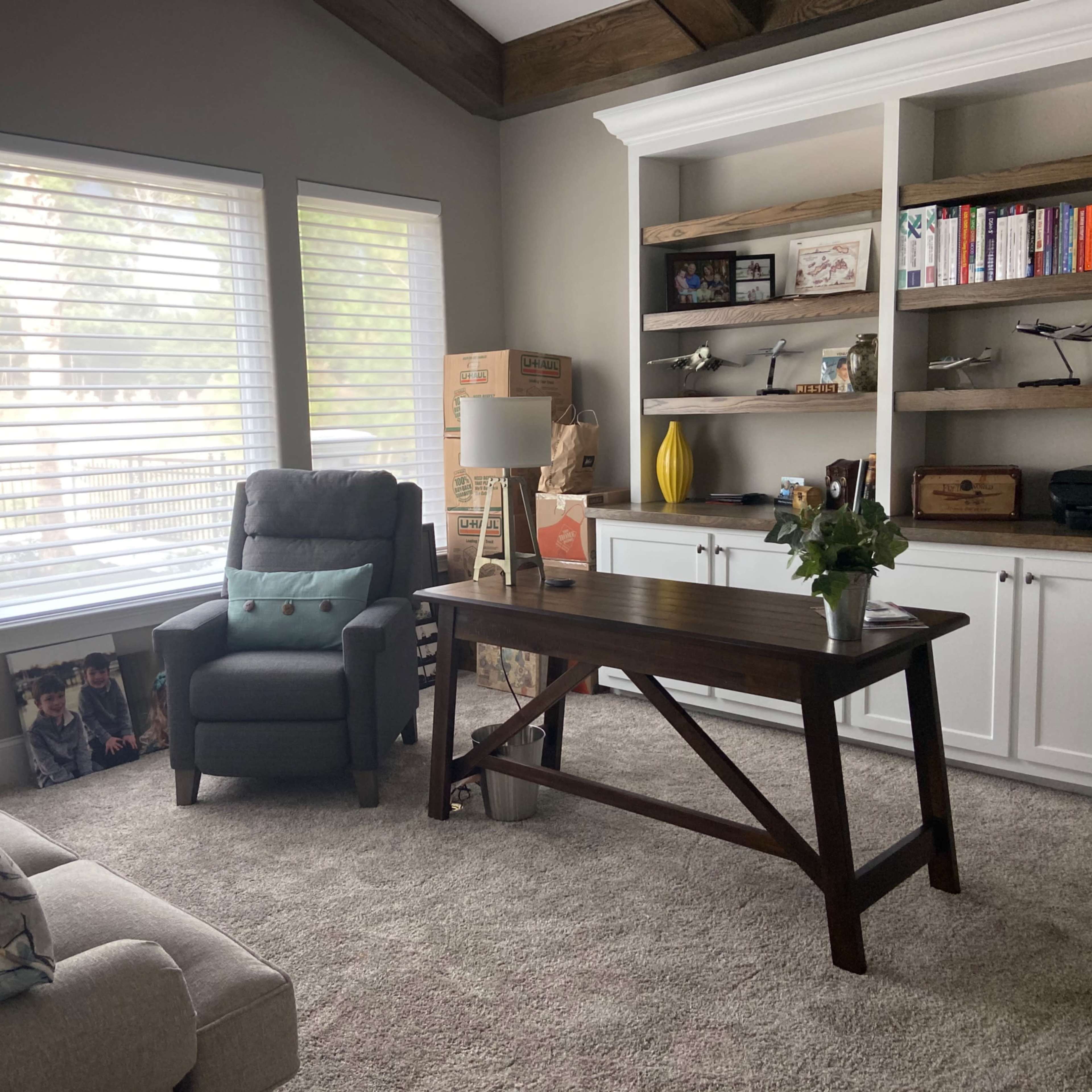 A cozy home office with a wooden desk, armchair, bookshelves filled with items, and large windows letting in natural light.