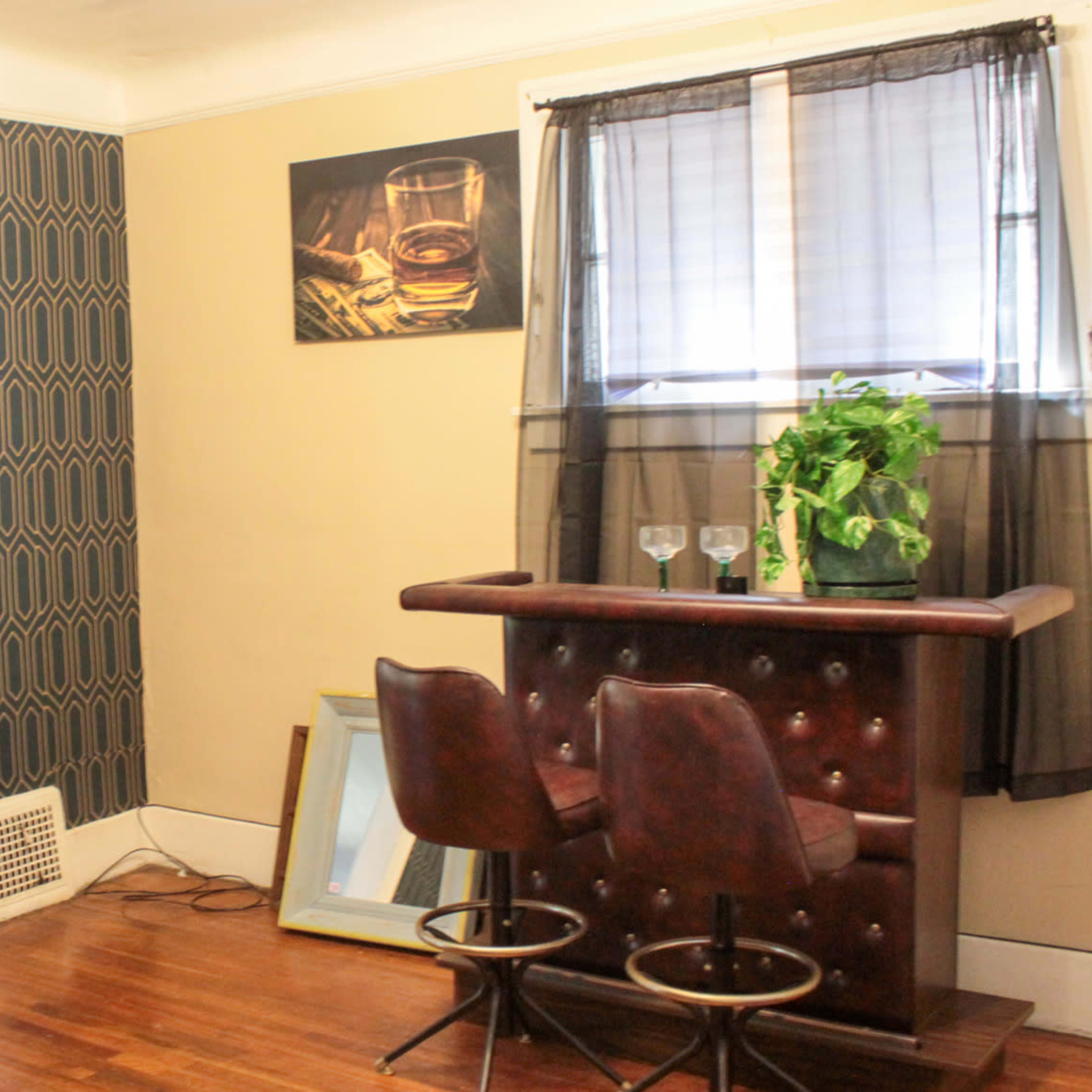A room with a small bar area featuring two stools, a potted plant, and a view into a separate living space with a couch and decorative pillows.