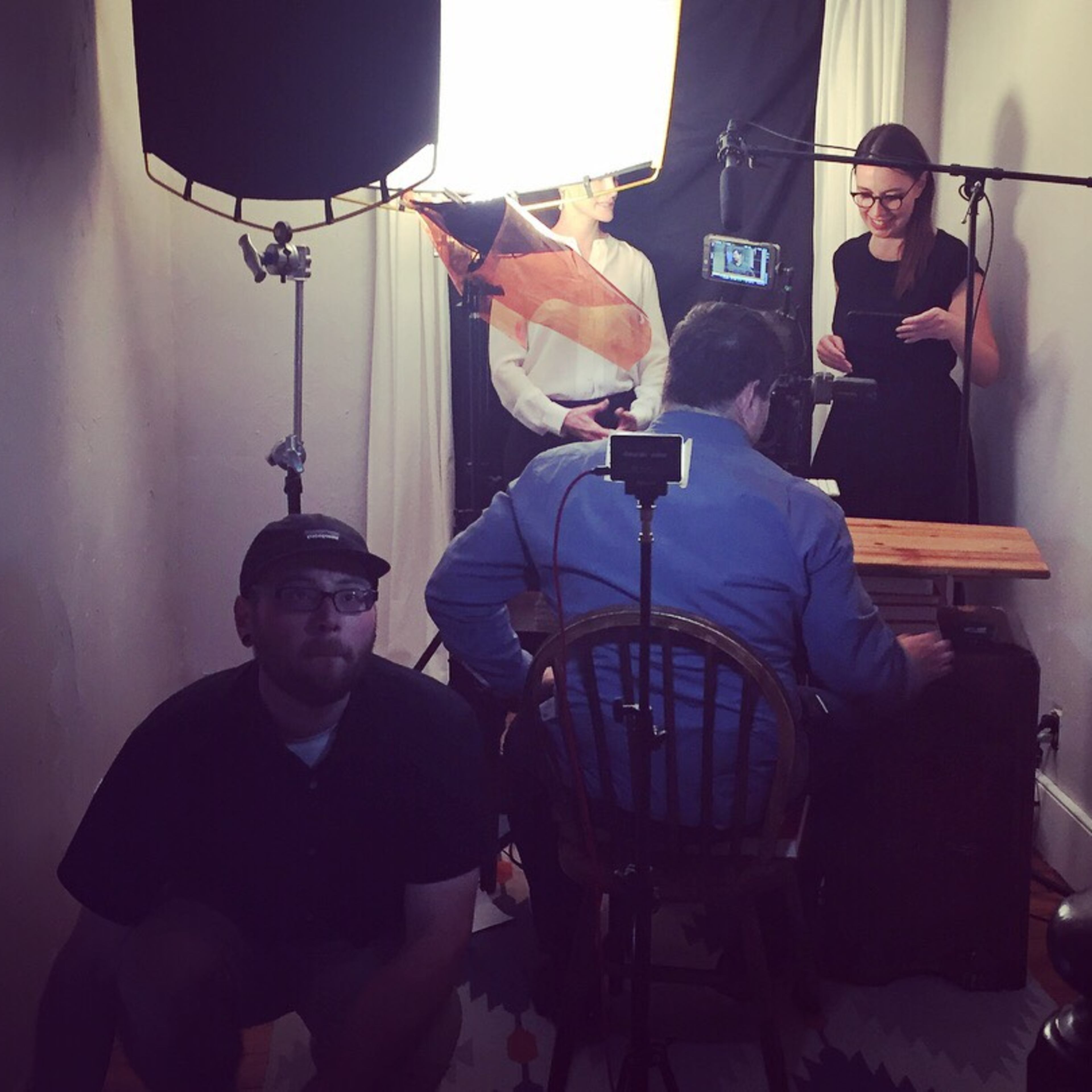 A small film crew is setting up equipment in a room to shoot a scene with a man seated at a table and a woman standing nearby, while another crew member adjusts a light.