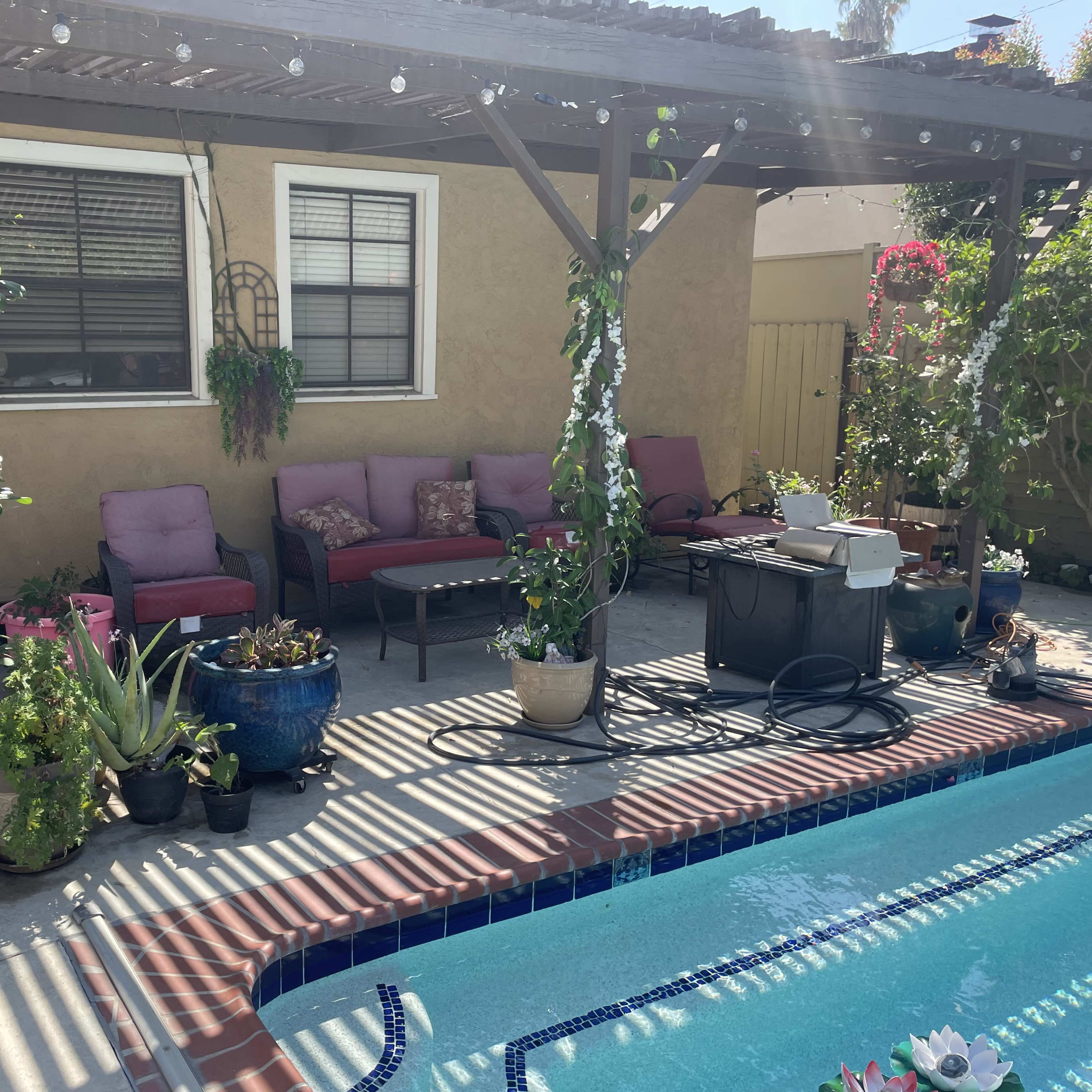 The scene features a backyard patio with a swimming pool, adorned with potted plants and seating arranged under a wooden pergola.
