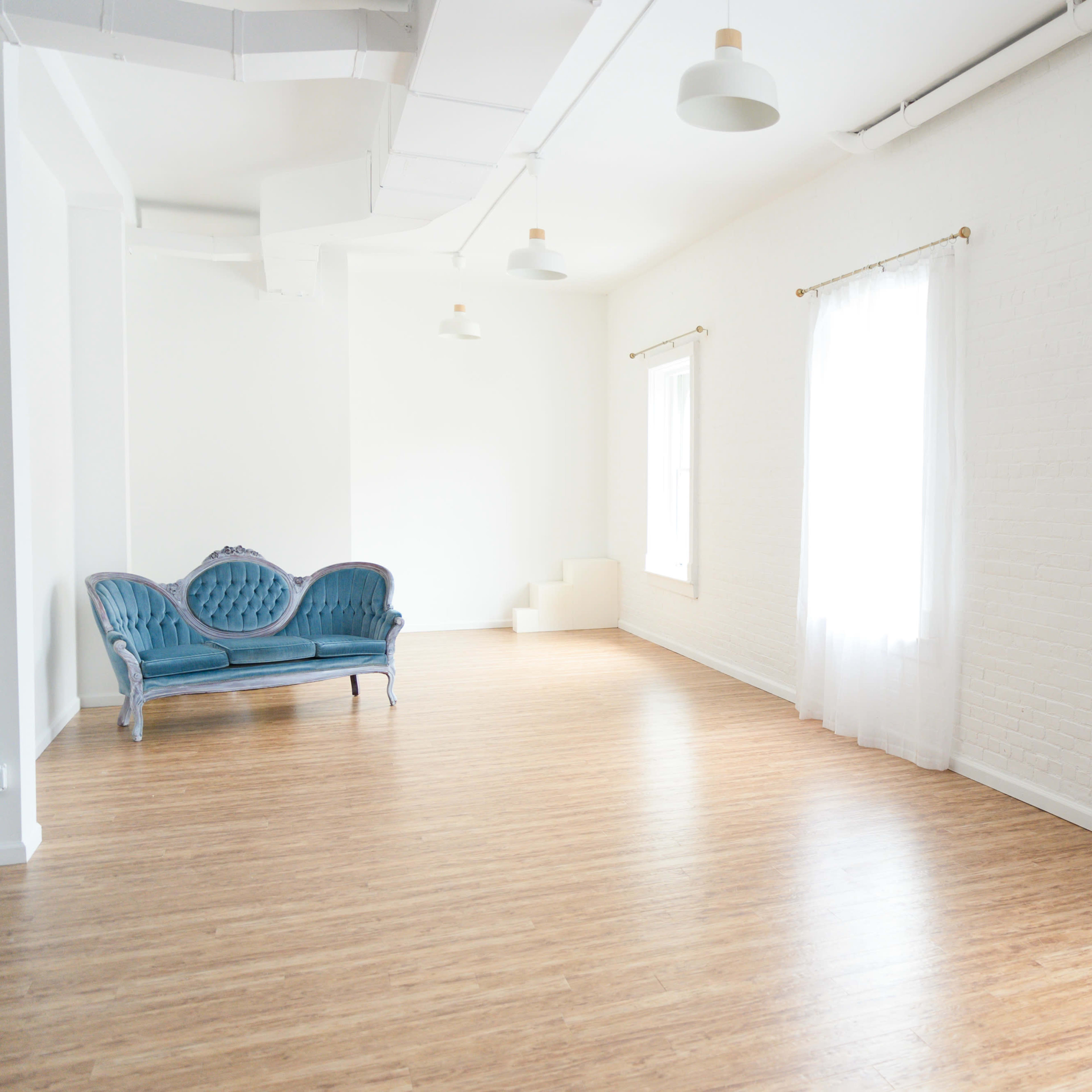 A bright, empty room features light wood flooring, white walls, two large windows with sheer curtains, and a blue velvet sofa against one wall.