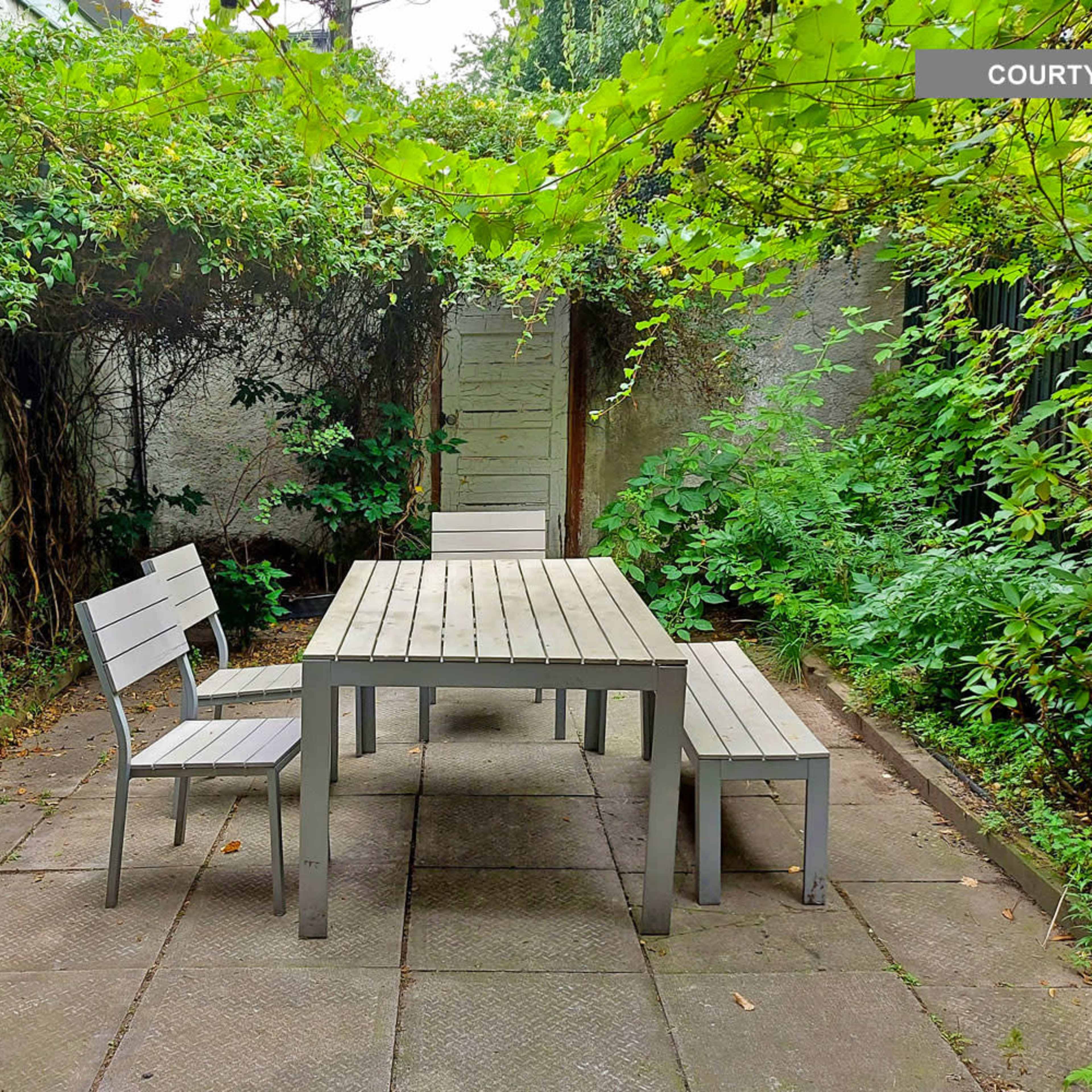 A small courtyard with a wooden table and two chairs surrounded by greenery and stone walls.
