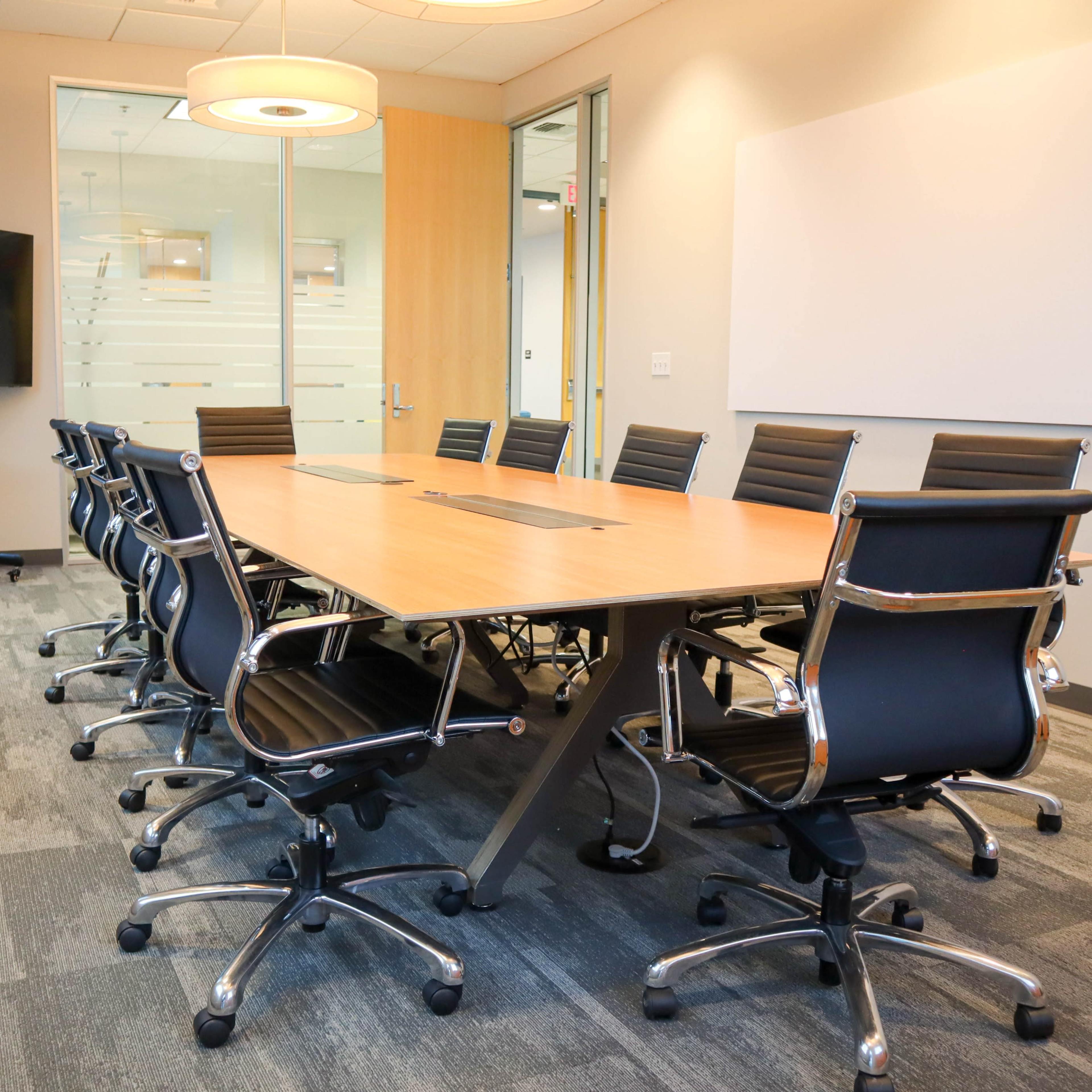 A conference room features a large wooden table surrounded by black rolling chairs, with a flat-screen TV and a whiteboard visible in the background.