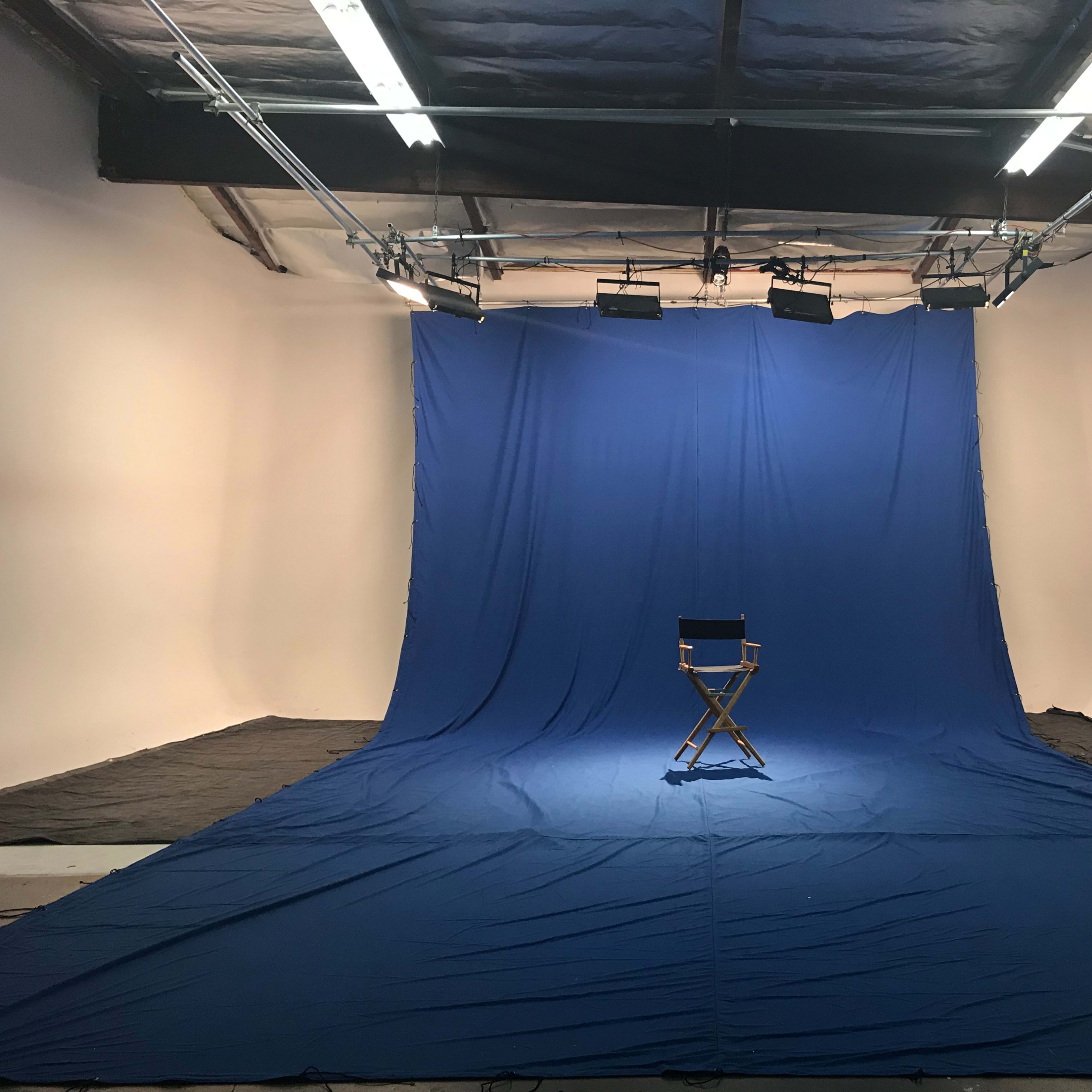 A blue backdrop covers the floor and walls of a studio, with a single director's chair positioned in the center under overhead lights.