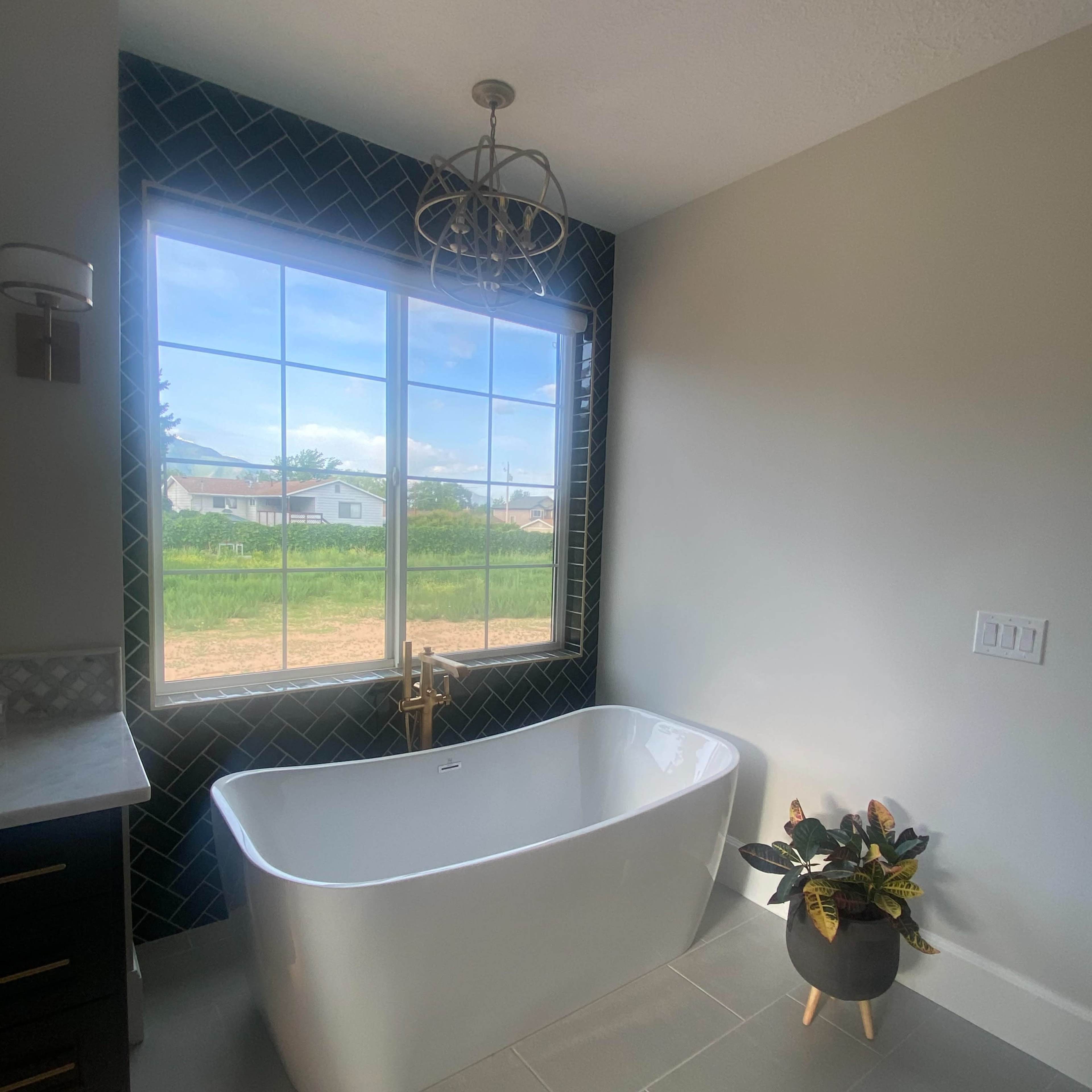 A modern bathroom features a freestanding tub beneath a large window, with dark tile accents and a potted plant nearby.