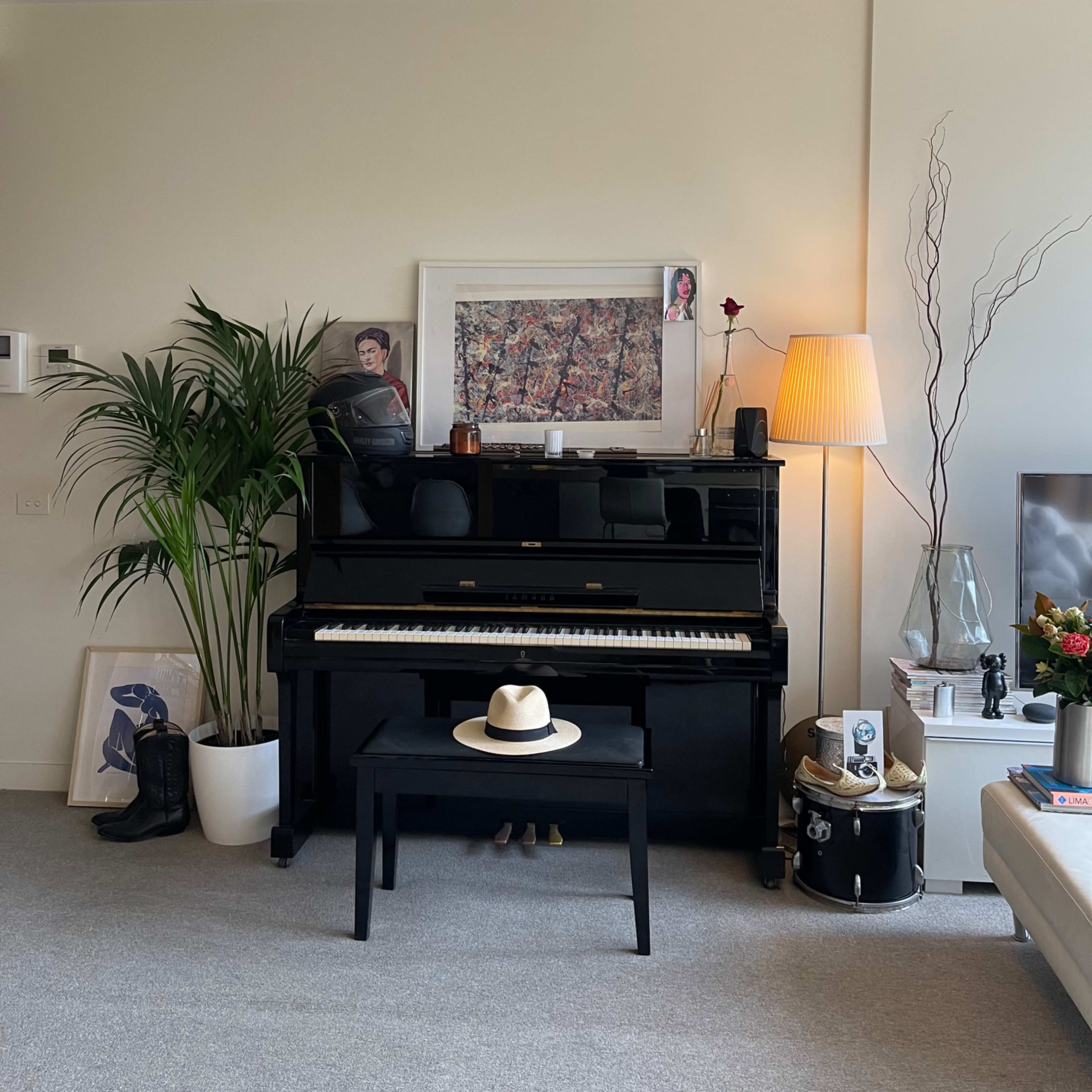 A living room with a black piano, a wooden bench, a hat on the bench, various plants, and artwork on the walls.
