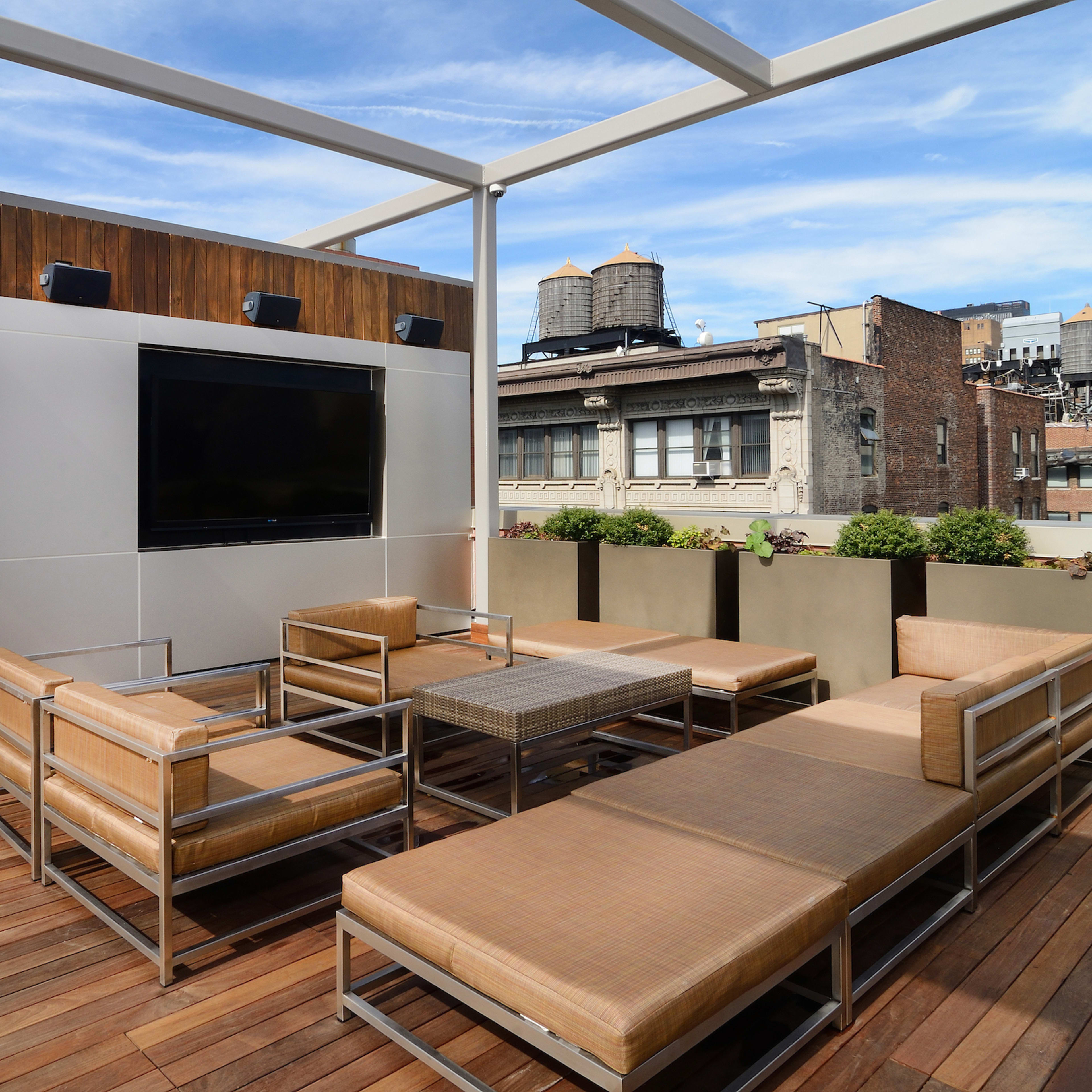 A rooftop terrace with metal-framed seating, a coffee table, and a large wall-mounted television, surrounded by potted plants and a view of city buildings.