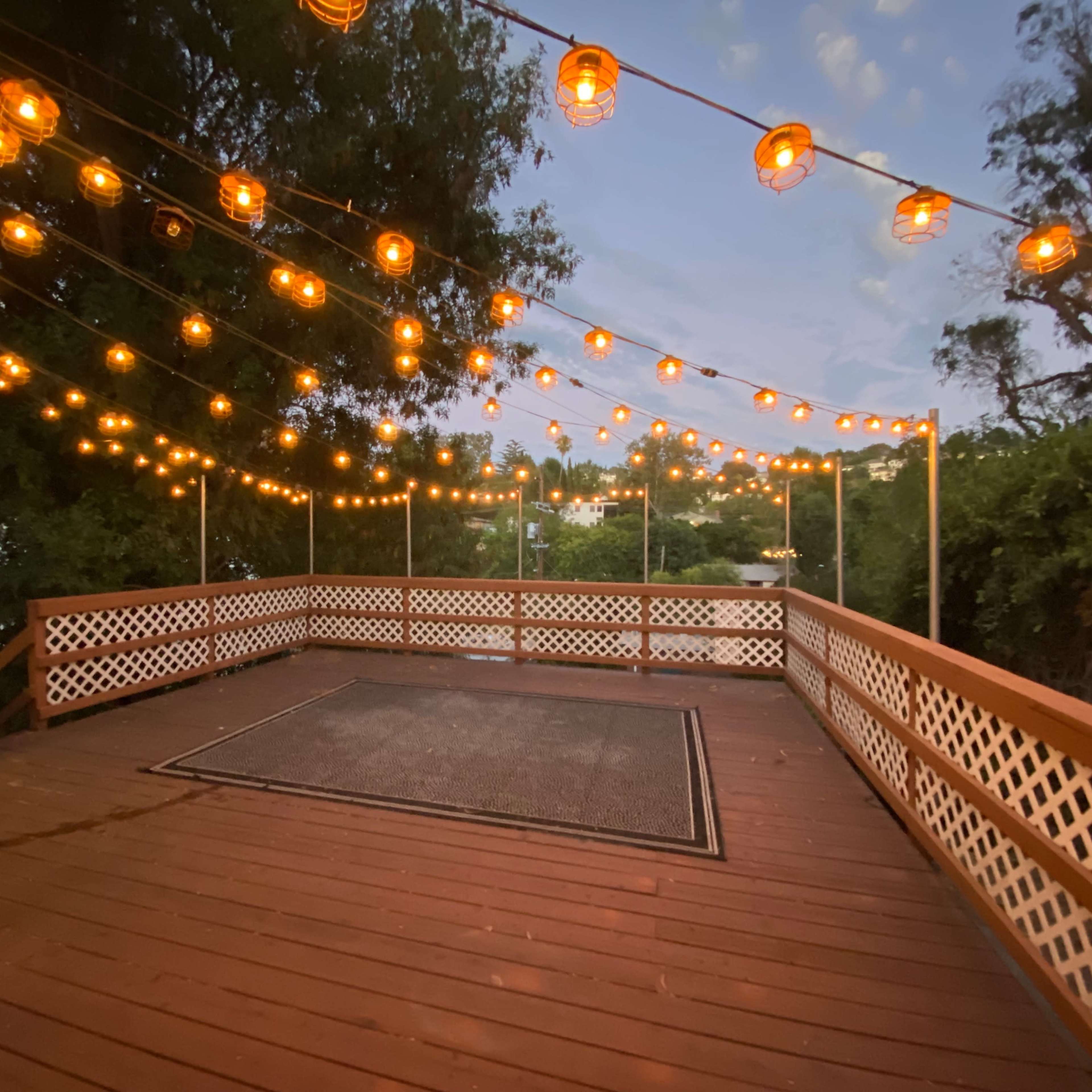 A wooden deck is illuminated by string lights, surrounded by greenery and overlooking a body of water at dusk.