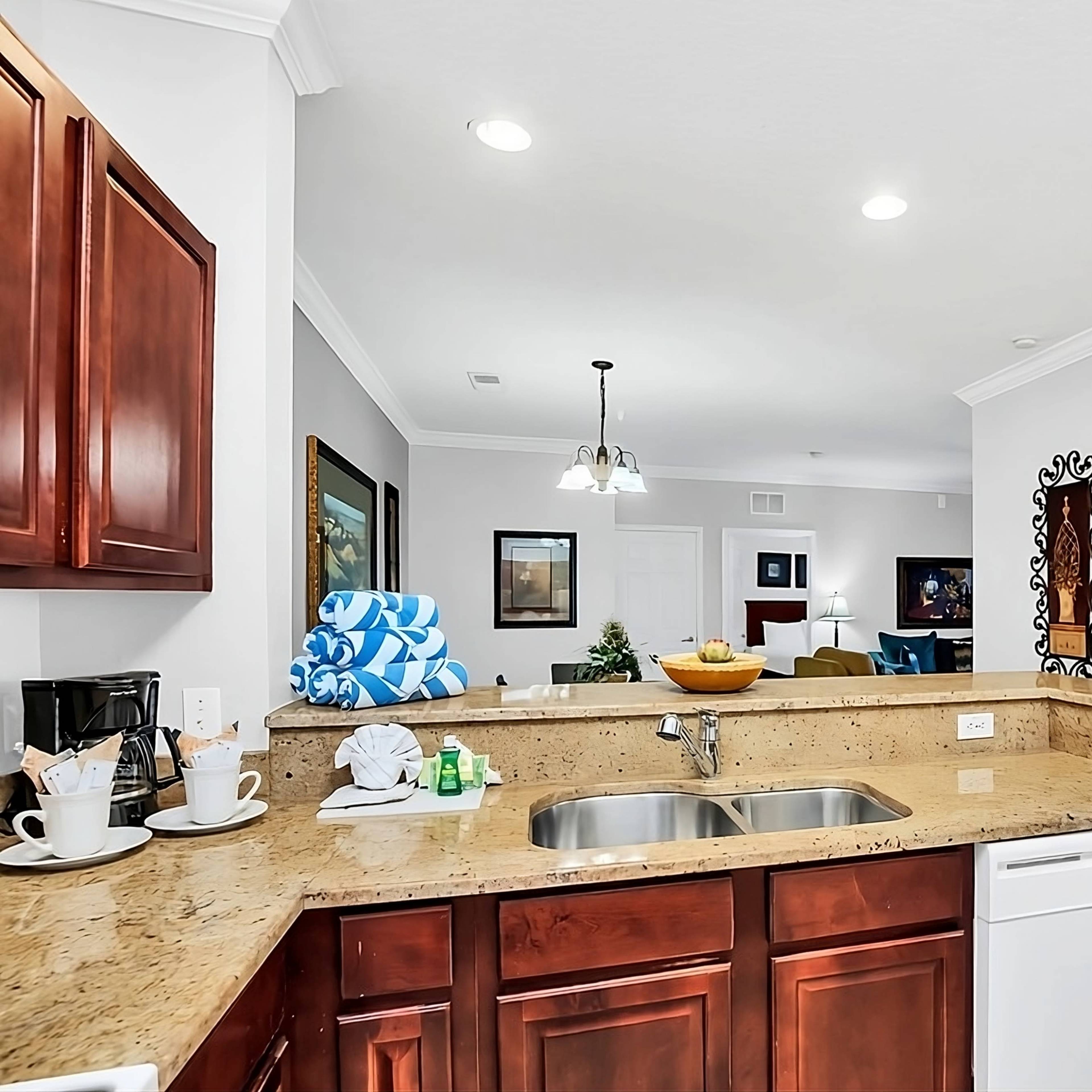 A kitchen area with wooden cabinets, a granite countertop, a sink, and a modern dishwasher, leading to an open living space.
