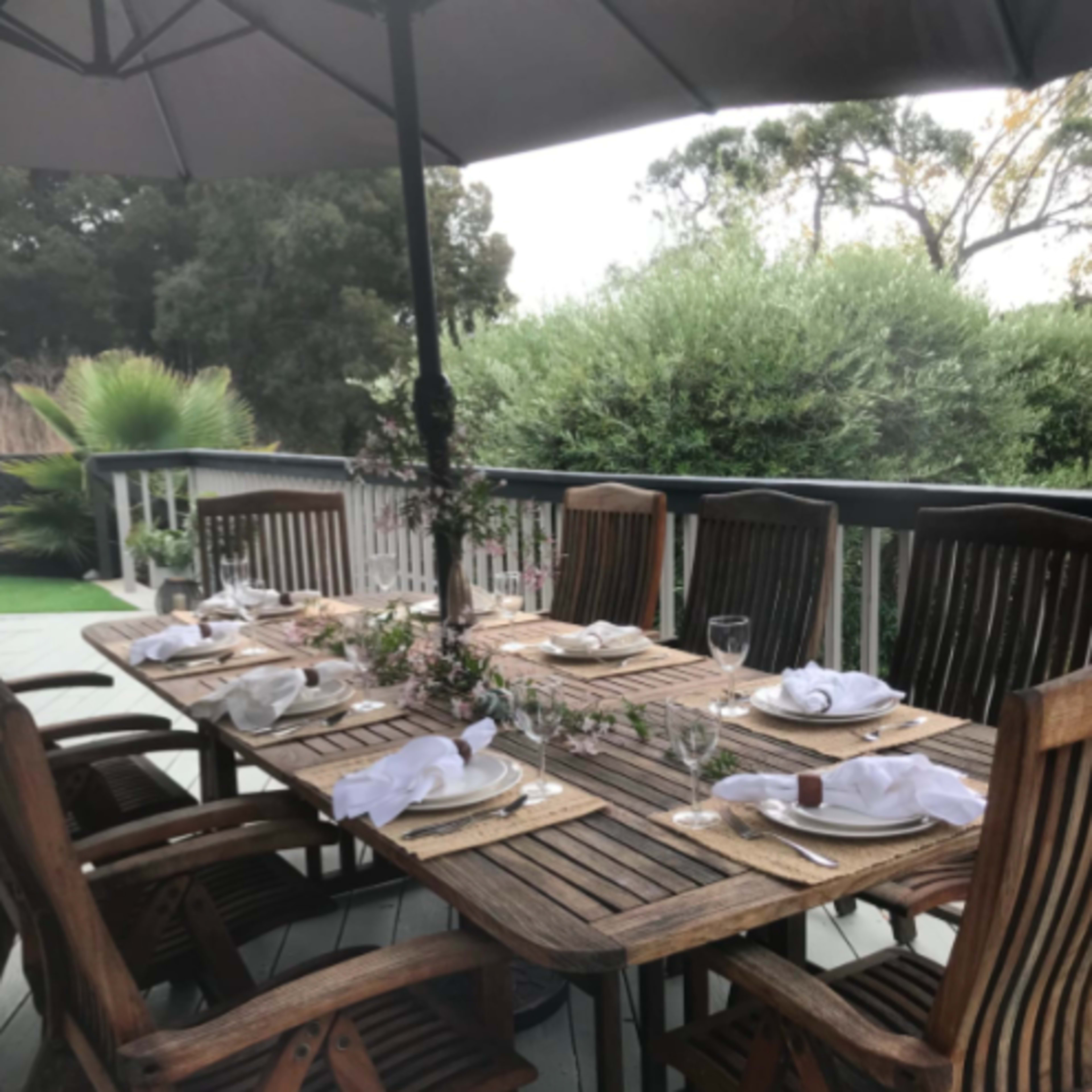 A wooden outdoor dining table is set with plates and glasses under a large umbrella on a deck surrounded by greenery.
