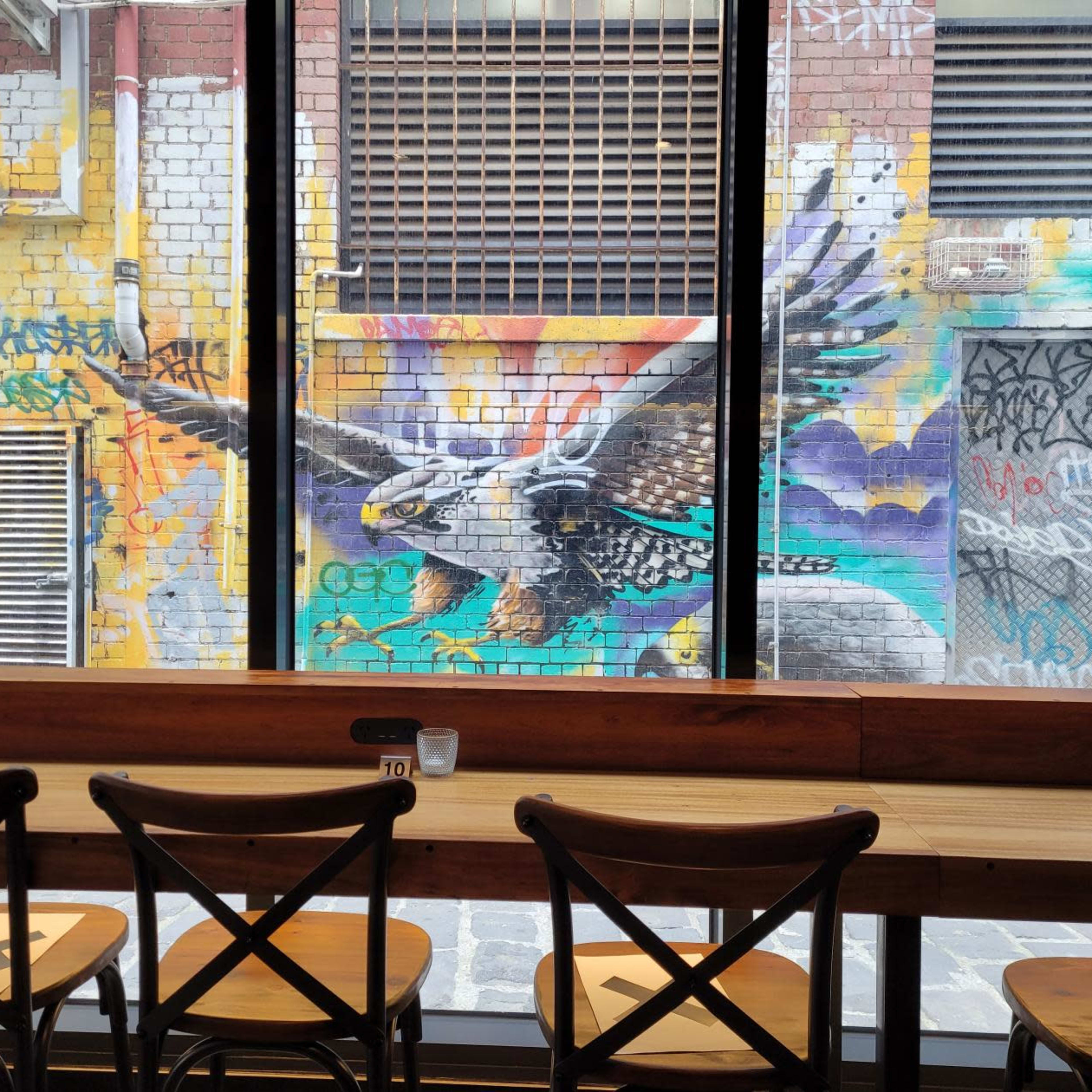 A mural of an eagle on a colorful brick wall, viewed through large windows from inside a café with wooden seating.