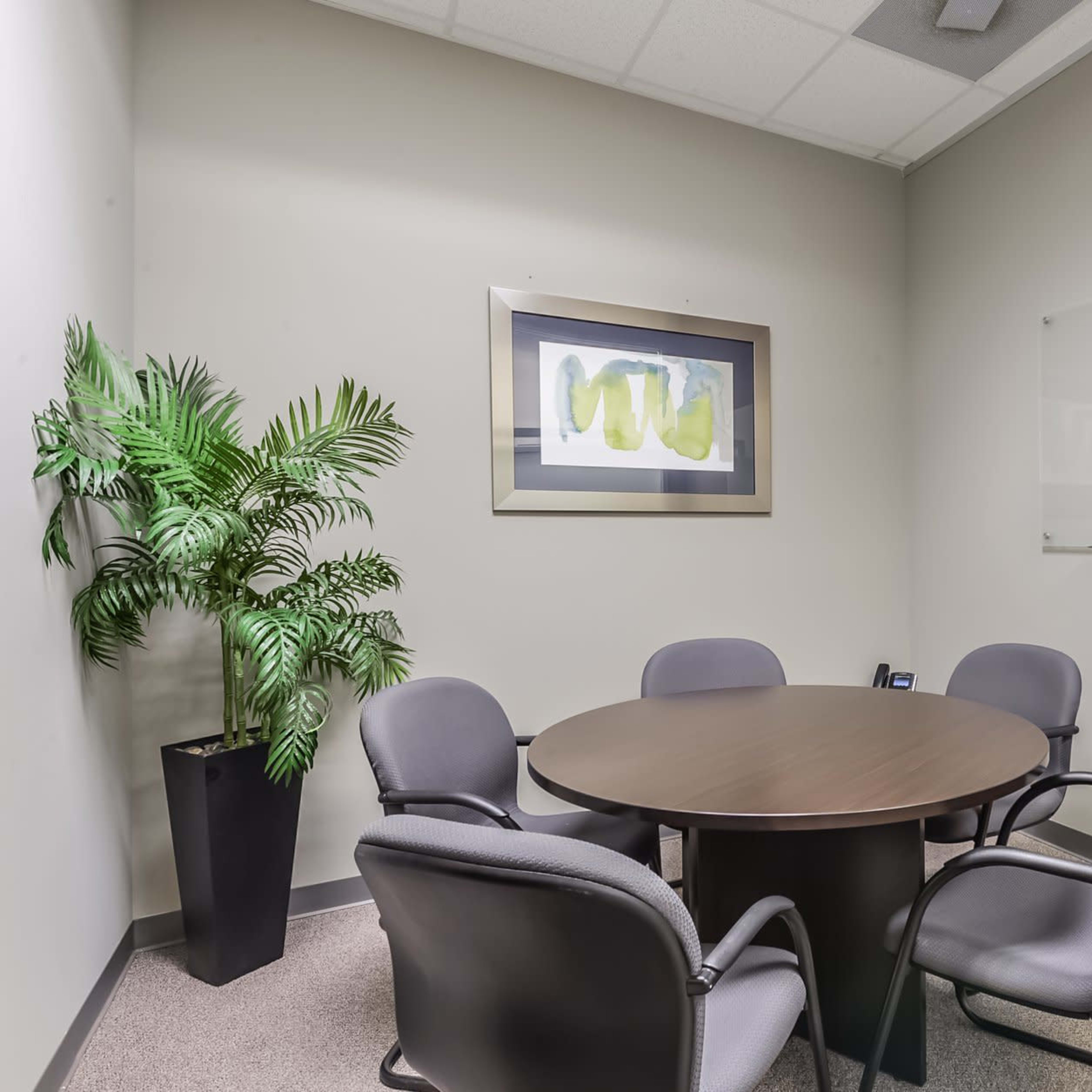 A small conference room features a round table surrounded by four chairs, a plant in the corner, and a whiteboard on the wall.