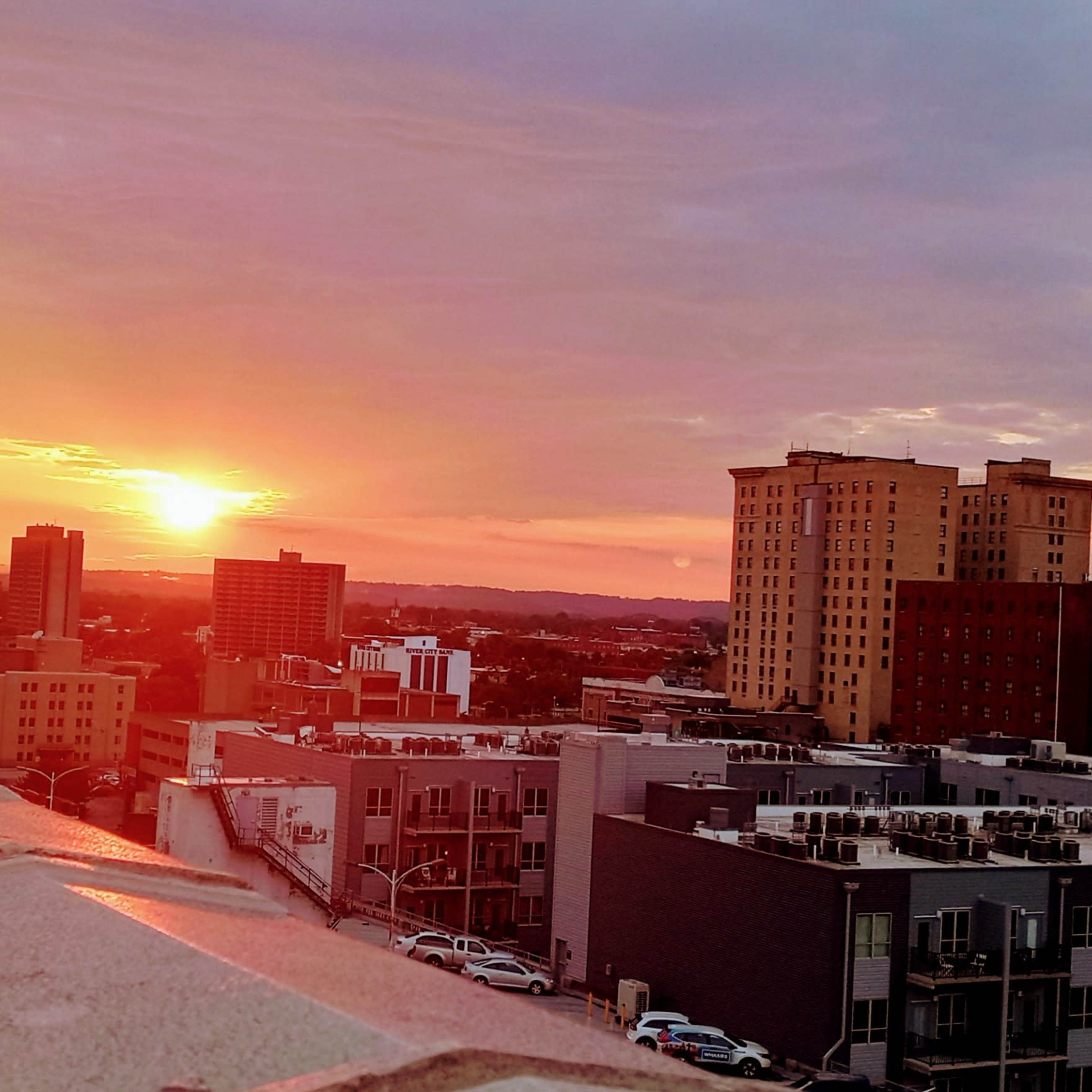 A sunset over a city skyline, with buildings and rooftops visible in the foreground.
