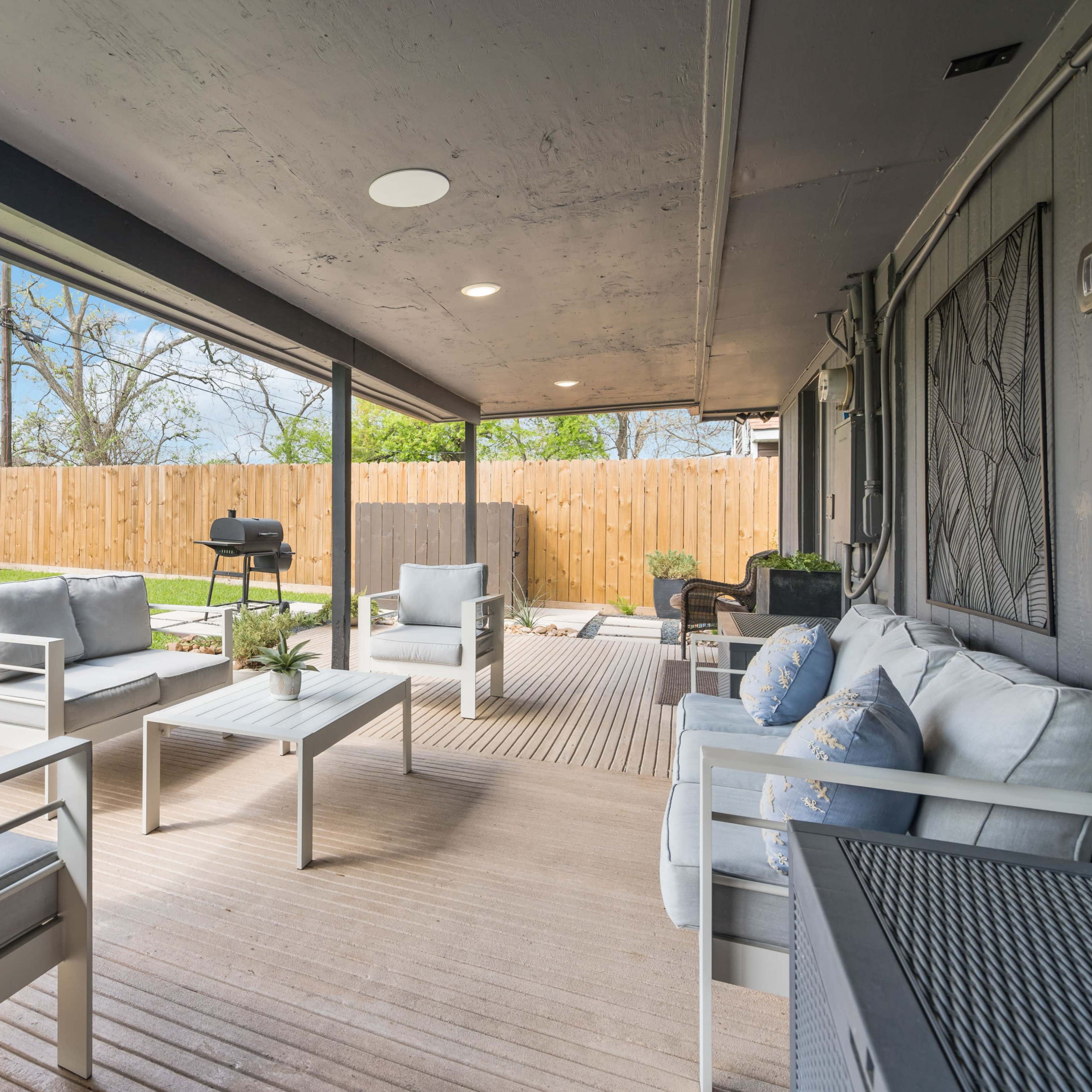 A covered patio with white furniture and a grill, surrounded by a grassy yard and wooden fence.